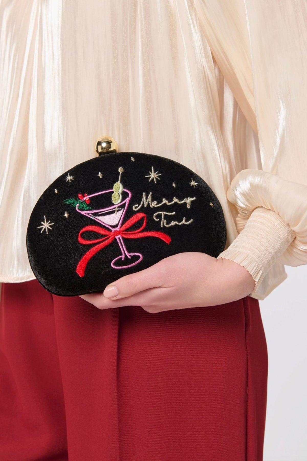 A person in a cream pleated blouse and red skirt holds a black velvet clutch decorated with a pink martini glass, red bow, gold stars, and the words 'Merry Time'.