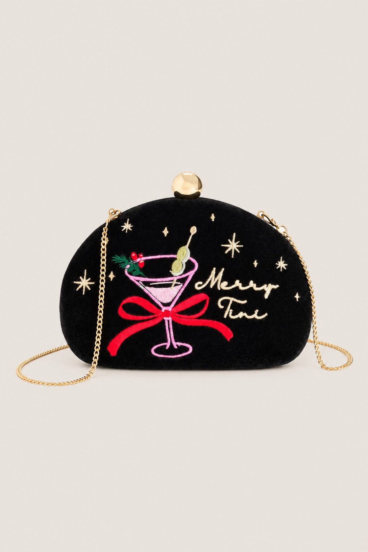 A black velvet crescent-shaped clutch bag with a gold-tone chain strap and top clasp. The front features embroidered holiday-themed details including a pink martini glass with green olives, a red bow, gold stars, and the cursive text 'Merry Fini'.