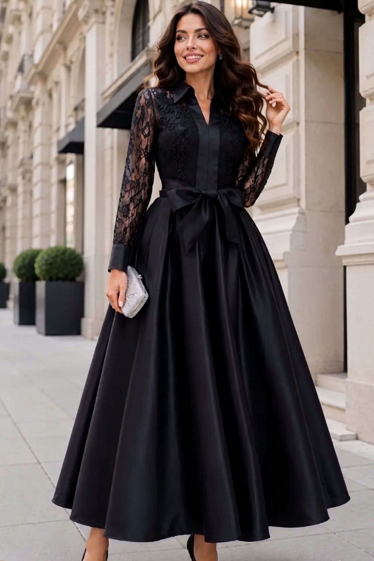 Woman wearing an elegant black maxi dress with sheer lace sleeves, satin bow waist and full skirt, styled with a silver clutch.