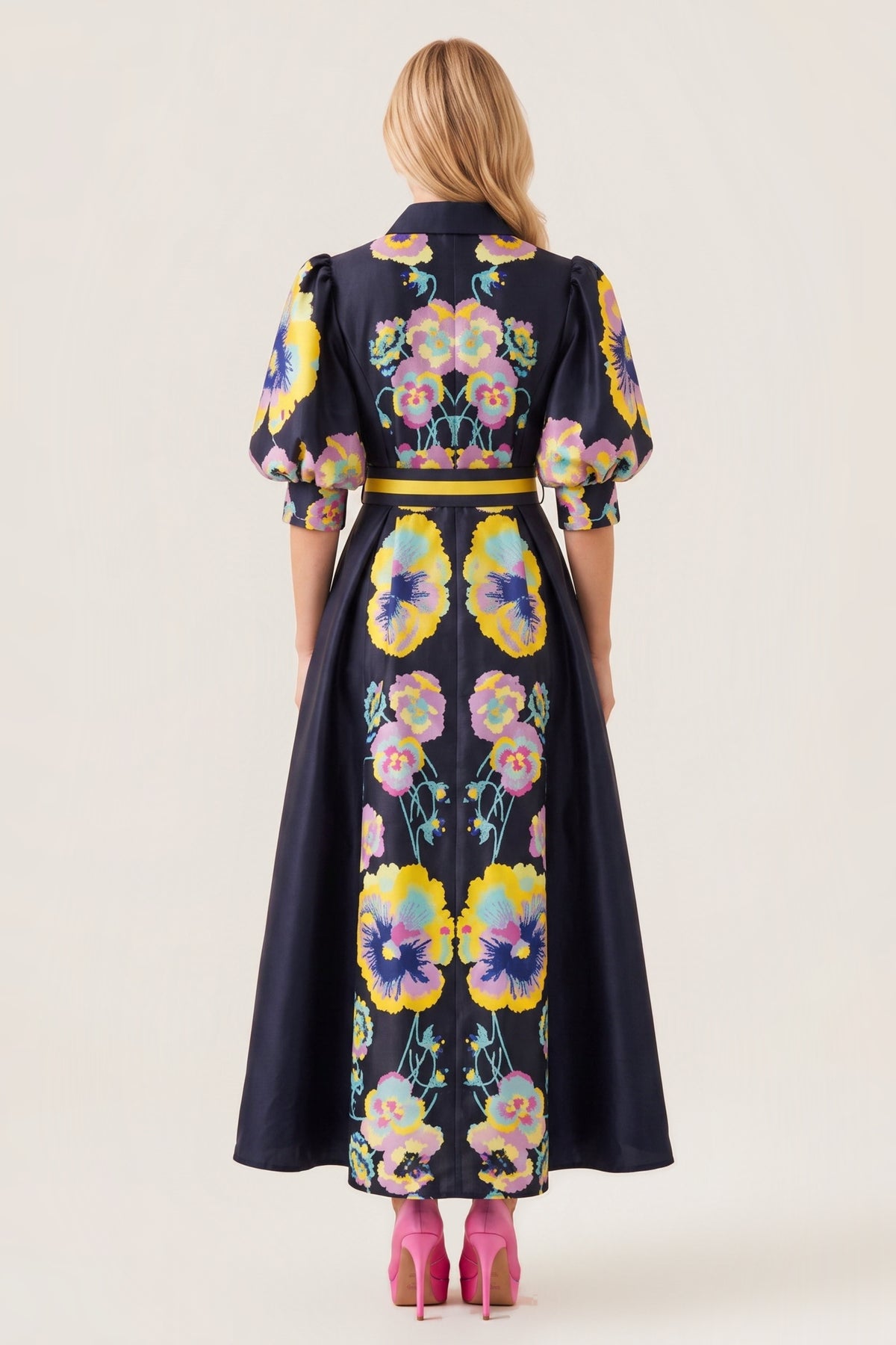 A woman viewed from the back wearing a long midnight dress with a colorful floral print and puff sleeves, paired with pink heels, against a plain beige studio background.