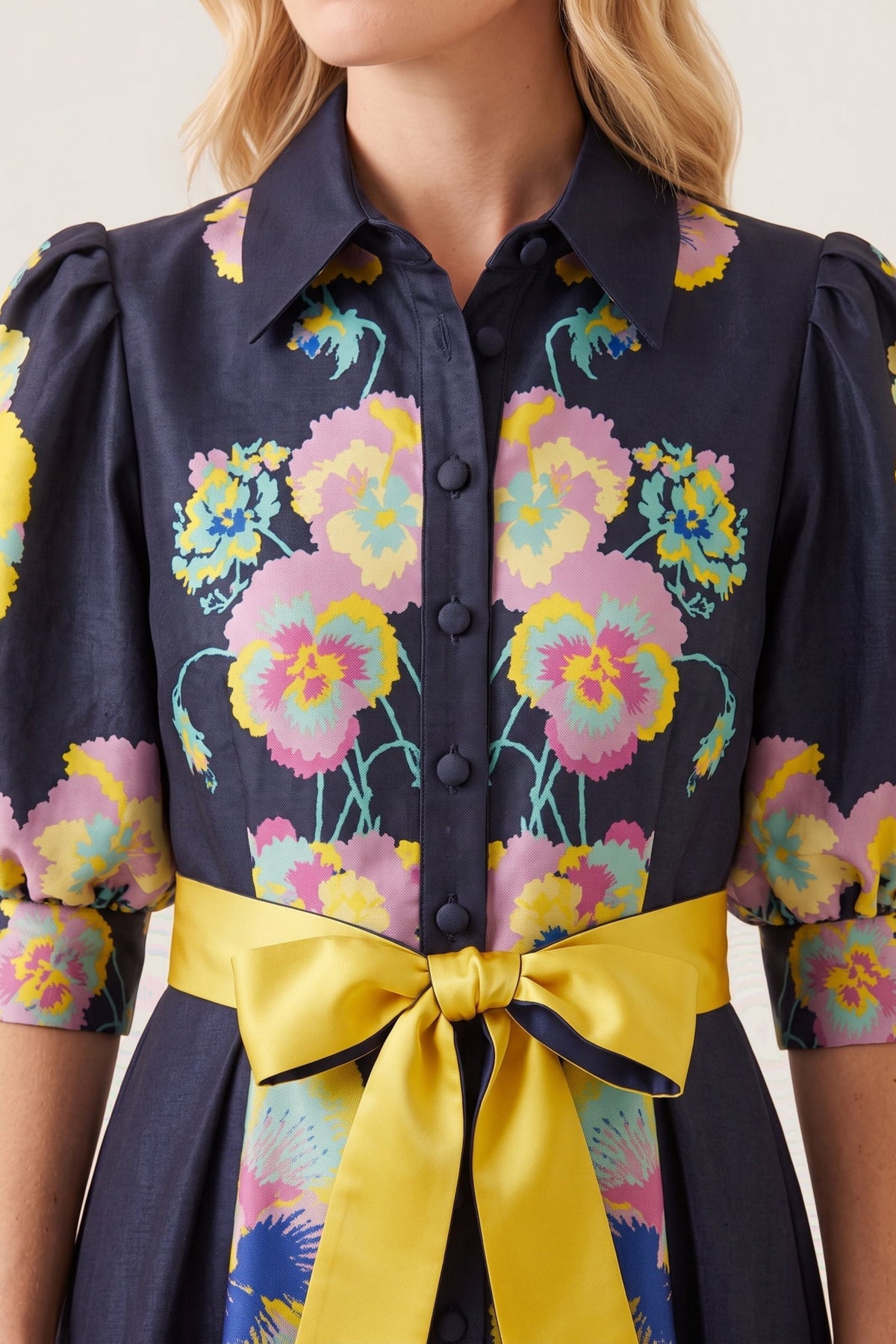 A close-up of a model wearing a midnight button-up dress with a vibrant multicolor floral print and a wide yellow satin waist tie bow.
