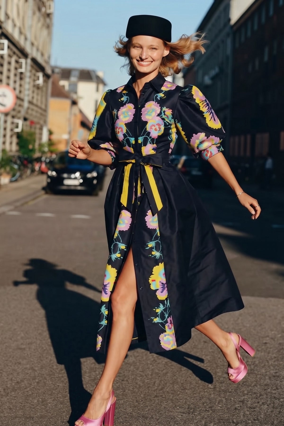 A woman wearing a black midi dress with a colorful floral print and a matching black hat, running on a city street.
