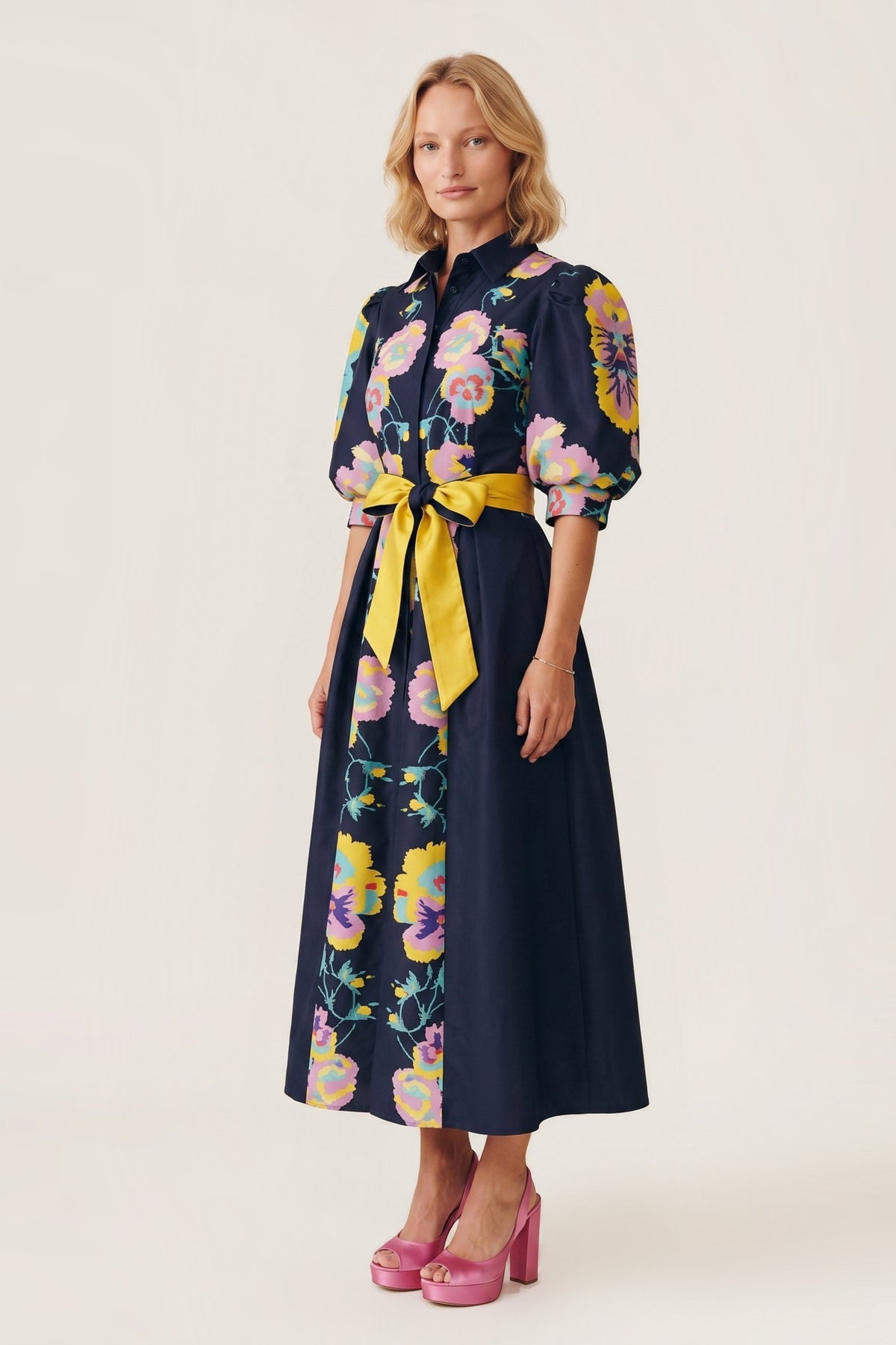 A woman modeling a midnight  midi dress with a colorful floral print and a bright yellow waist tie, paired with pink high heels, against a plain beige studio background.