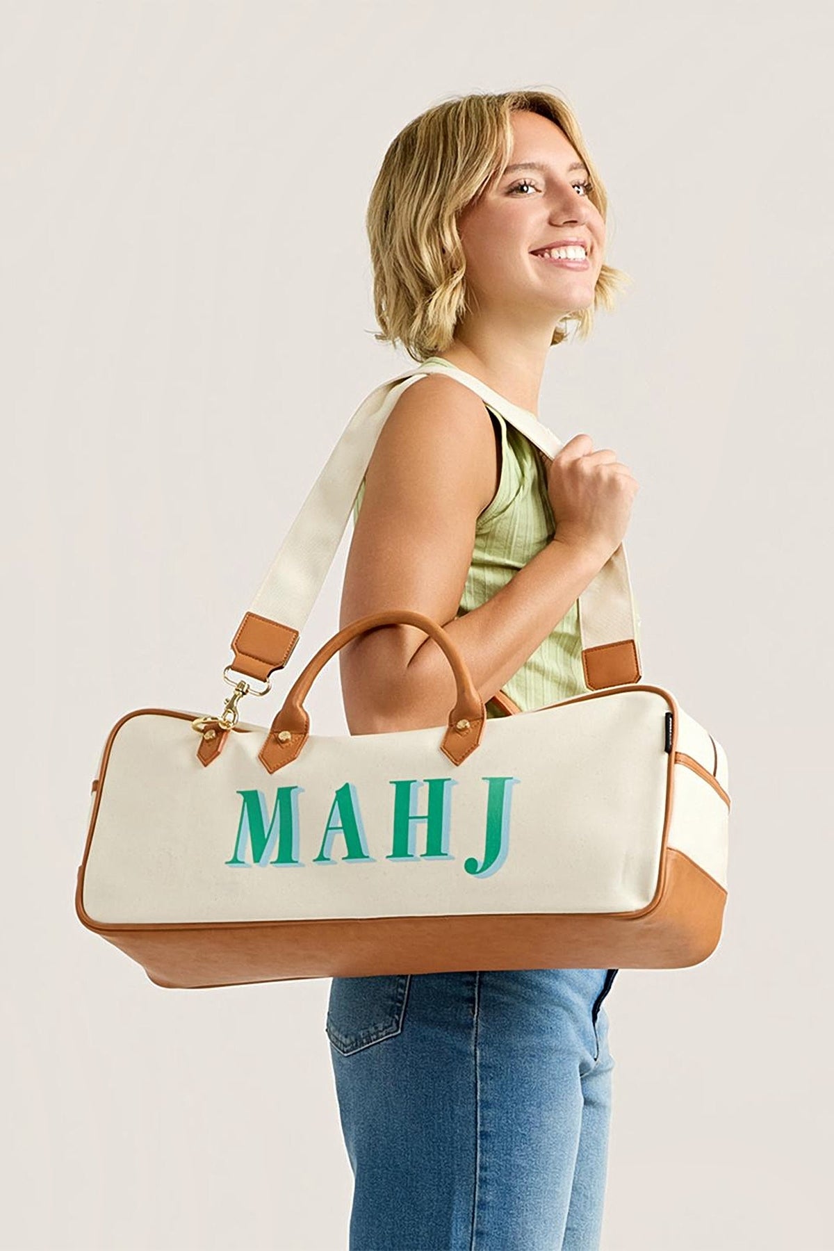 A woman carrying the Mint Margo mahjong tote on her shoulder by its canvas strap, mint MAHJ lettering facing out.