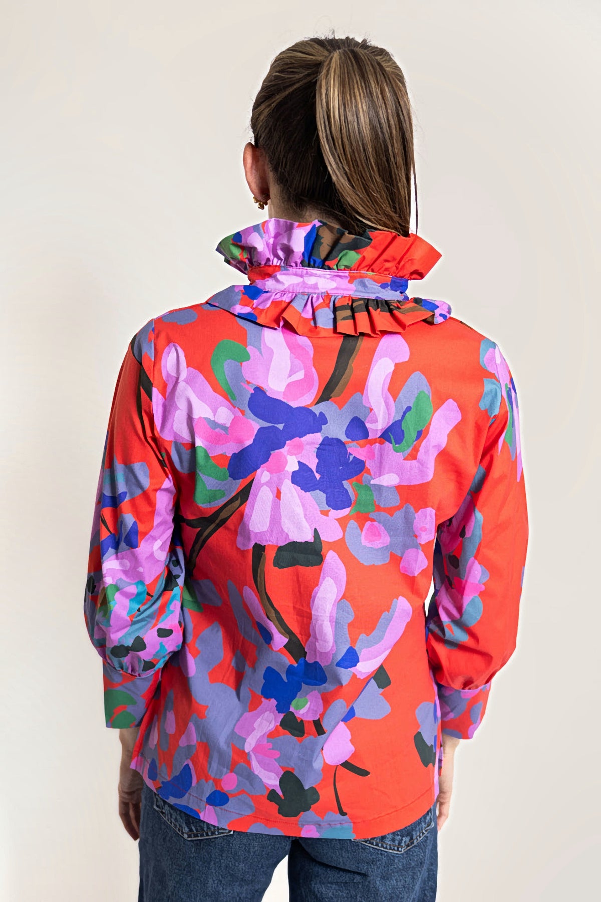 A woman shown from the back wearing a bright orange long-sleeve blouse with a large multicolor floral print in pink, purple, blue, and green, paired with blue jeans against a plain white background.