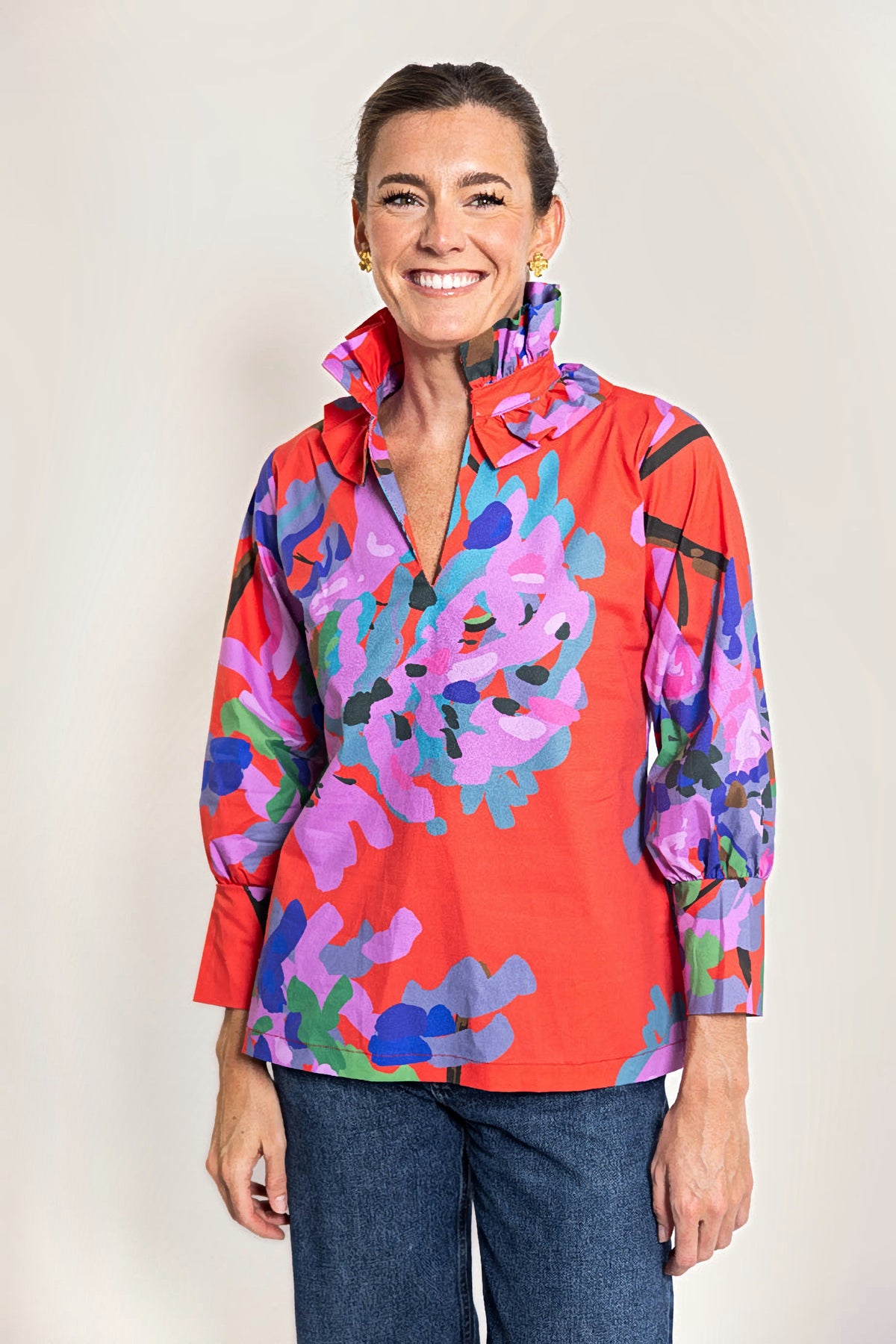 A woman wearing a bright red long-sleeve blouse with a bold abstract floral print in pink, purple, blue, and green, paired with dark blue jeans, against a plain white background.