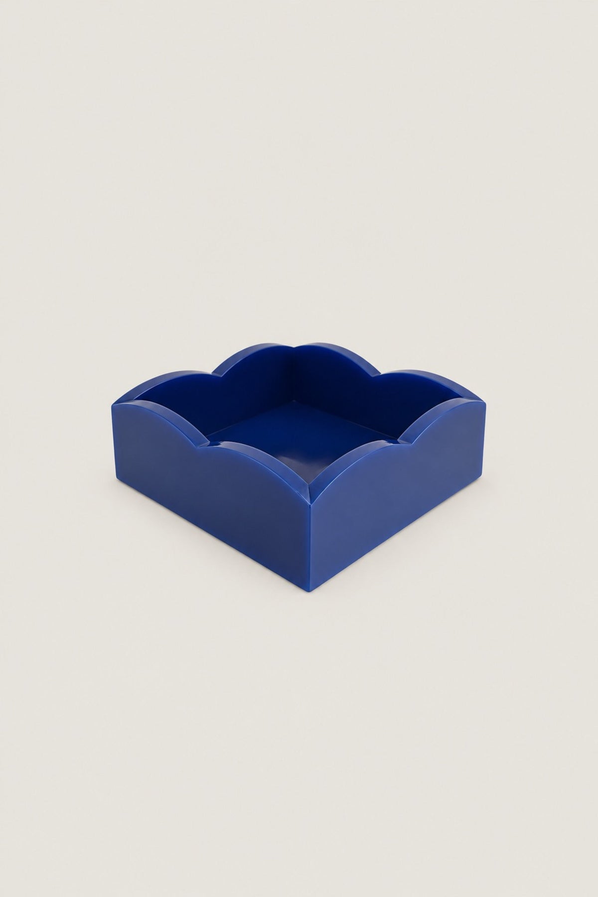 A small square tray with a scalloped or wavy rim, shown in a solid deep cobalt blue color. The tray is photographed on a clean, solid, light beige background with no other objects or people present.