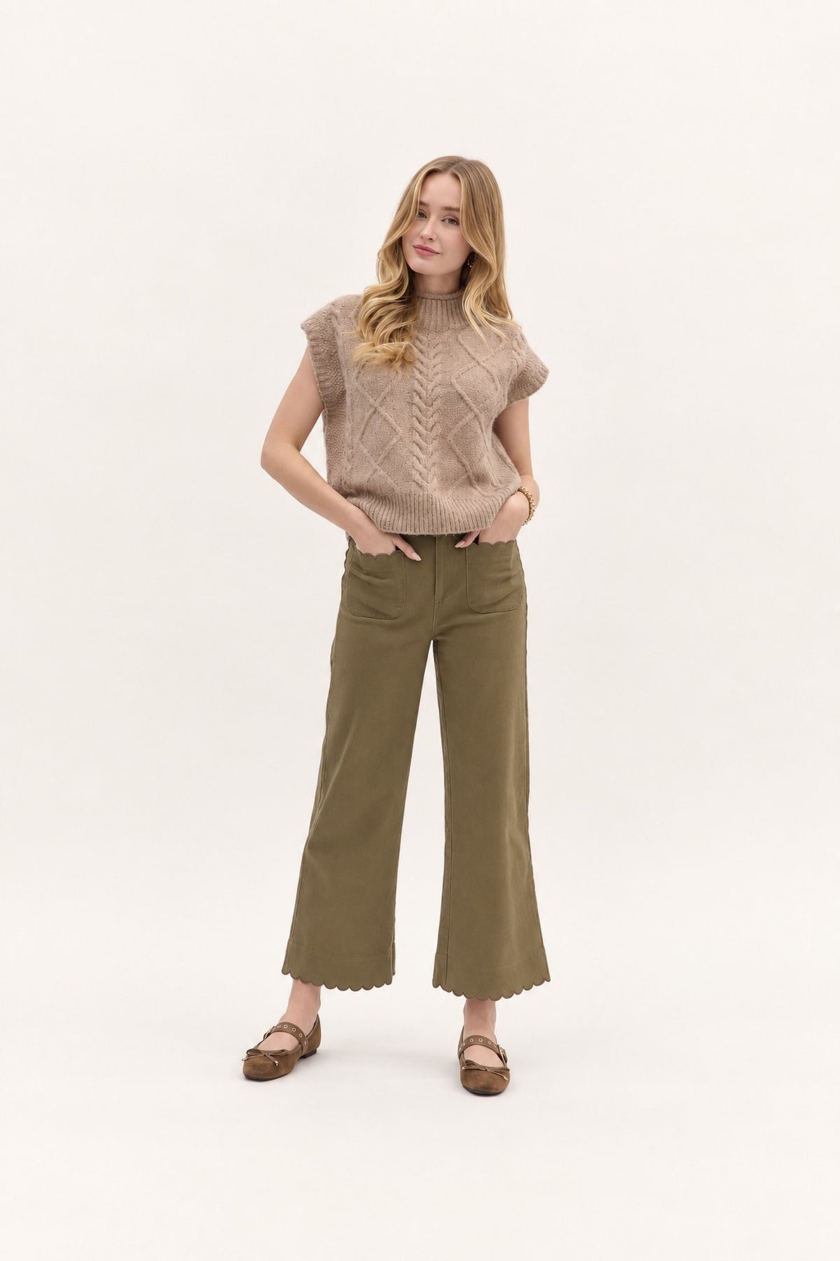 A woman modeling a beige cable-knit sleeveless turtleneck sweater vest and olive green wide-leg cropped pants with scalloped hems, paired with brown flat sandals, against a plain white studio background.