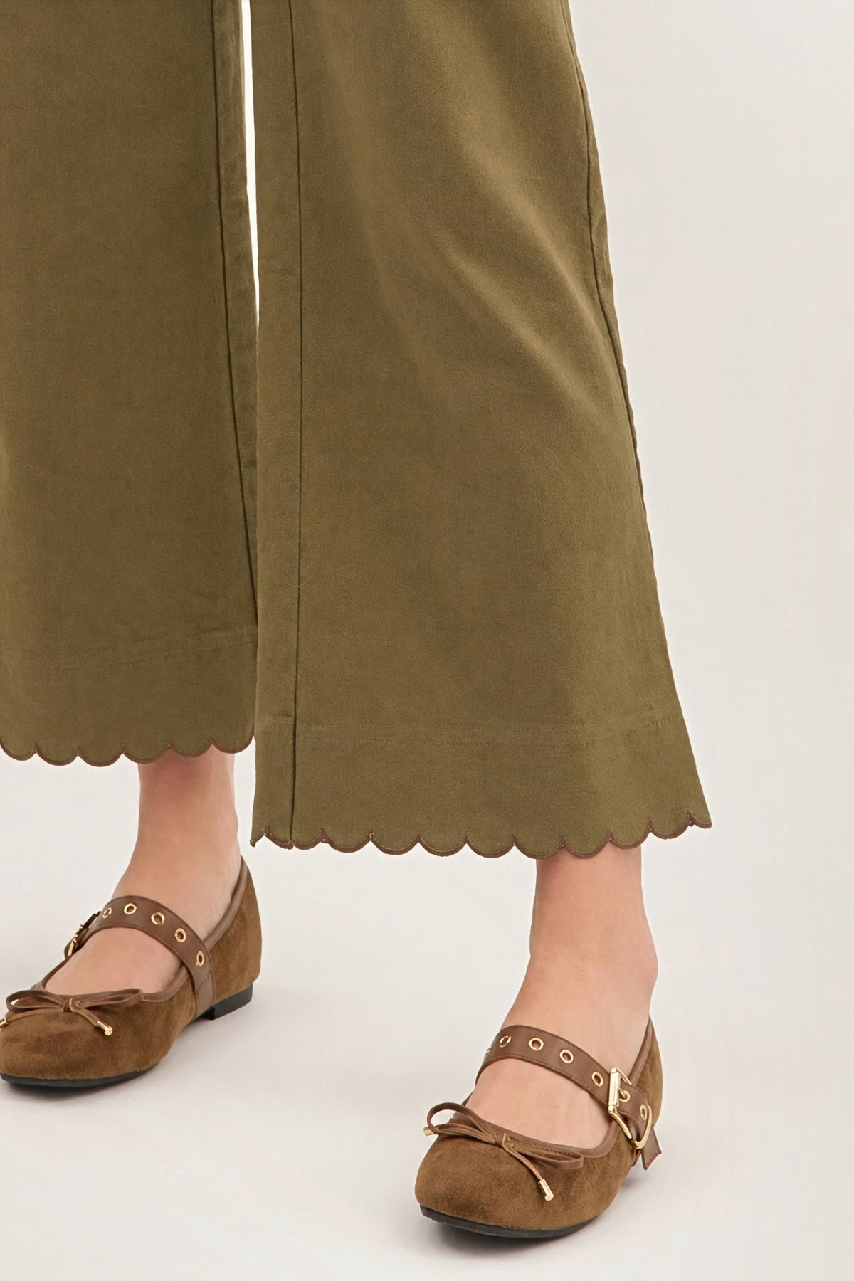 A close-up of a person wearing olive green wide-leg cropped pants with scalloped hems and brown suede Mary Jane flats with gold-tone eyelets and bows, shown against a plain light background.