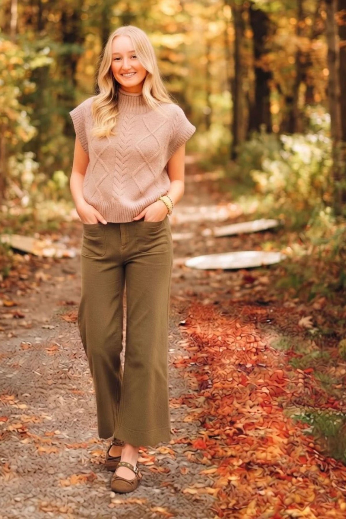 A woman stands on a leaf-strewn forest path wearing a sleeveless ribbed knit turtleneck sweater in a dusty blush pink and olive green wide-leg pants, paired with brown flat shoes.