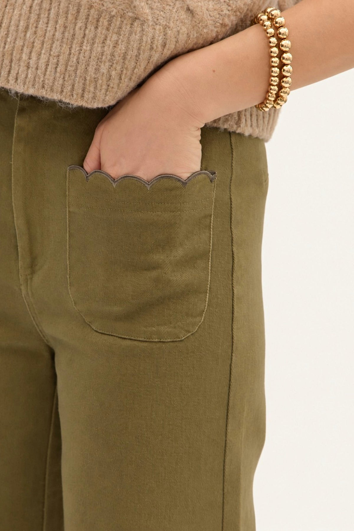 Close-up of a person wearing olive green trousers with a scalloped pocket edge, a beige ribbed sweater, and gold beaded bracelets against a light neutral background.