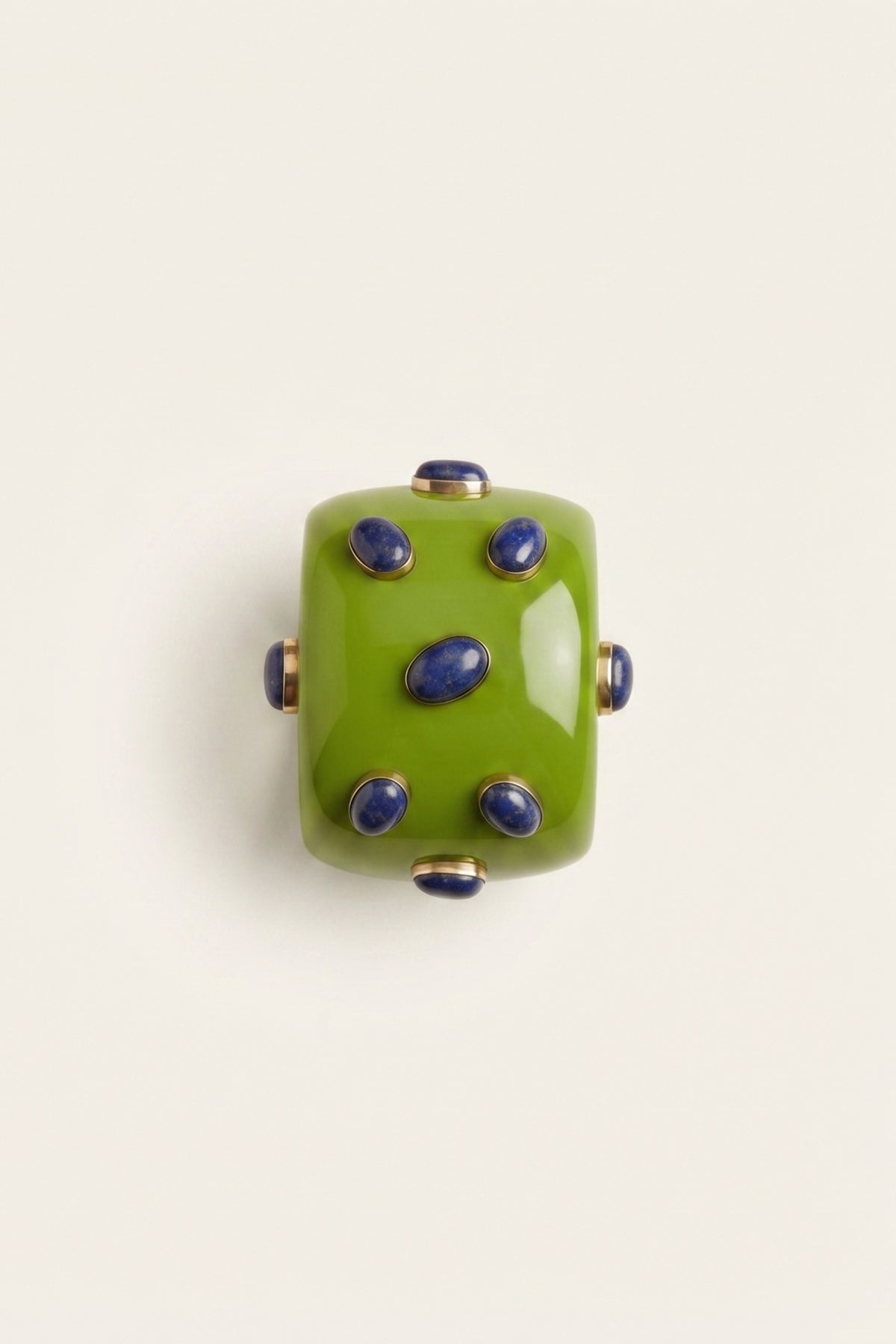 A square green glass bead with six blue cabochon stones set into a gold-toned metal frame, photographed from directly above on a light cream background.