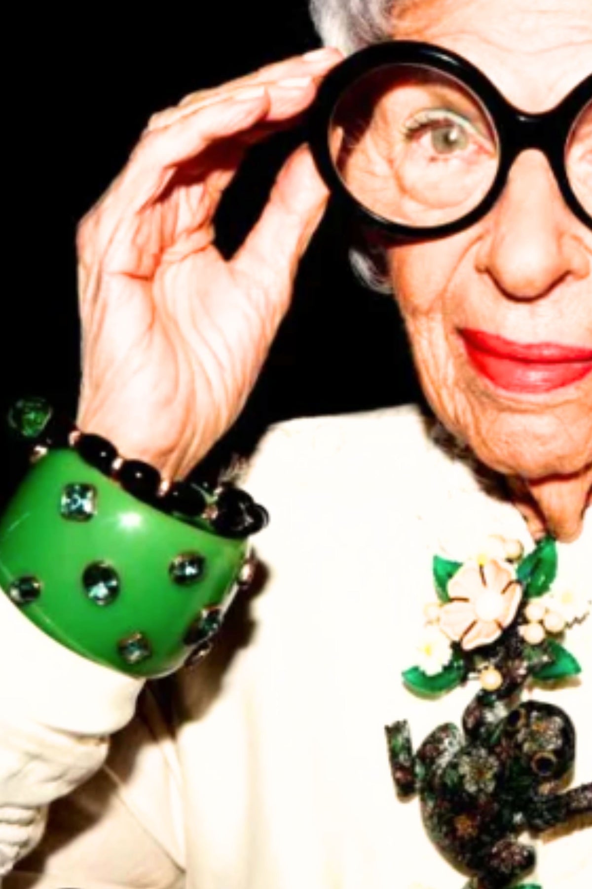 An elderly woman with white hair and glasses adjusting her frame, wearing a large green beaded cuff bracelet with black rhinestones and a white jacket with a green and white floral brooch, against a black background.