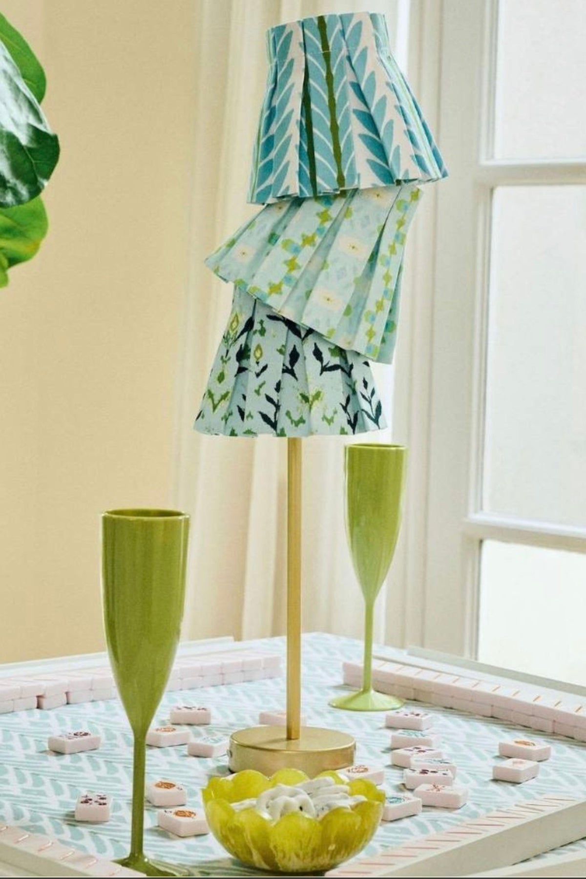 A decorative lamp with tiered fabric shades in teal, green, and white patterns, displayed on a table with matching green glassware and small pink decorative items in a bright indoor setting.