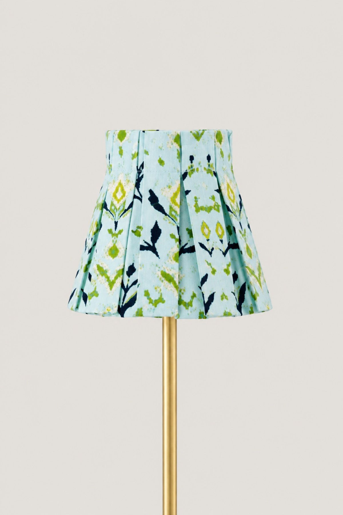 A pleated fabric lampshade with a light blue background and a green, yellow, and black botanical pattern, mounted on a gold lamp stand against a solid light beige background.