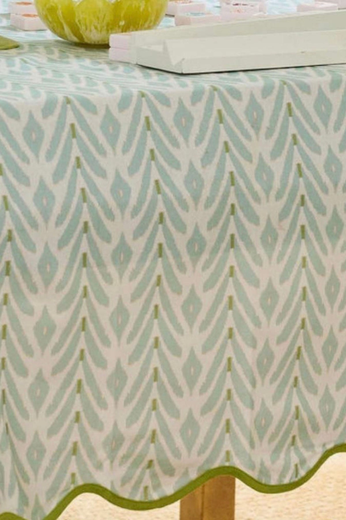A close-up of a tablecloth with a repeating vertical ikat-style pattern in pale aqua and olive green on an off-white background, with a scalloped green edge and part of a wooden table leg visible.