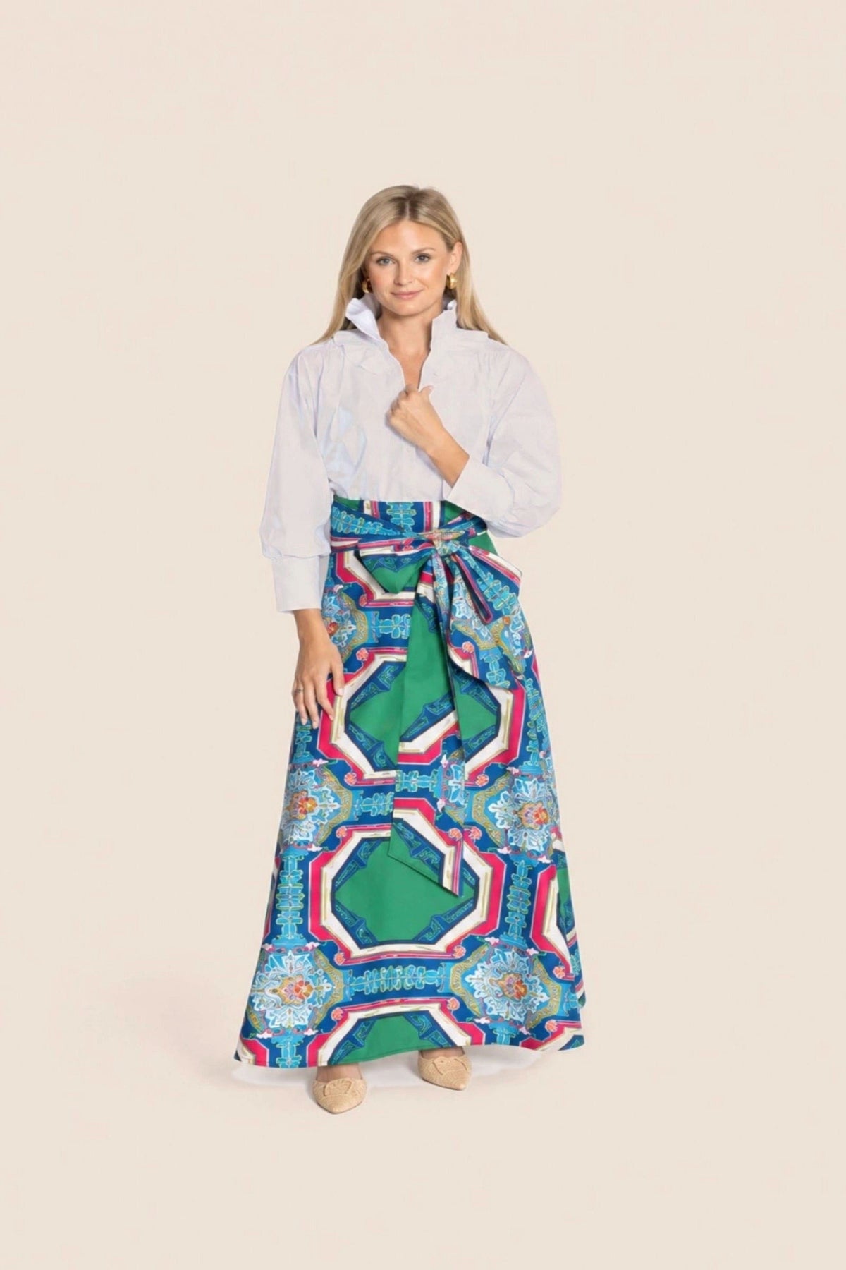 A woman modeling a white long-sleeve collared shirt tucked into a long, wide skirt with a bold geometric pattern in green, blue, pink, and white, standing against a solid light beige background.