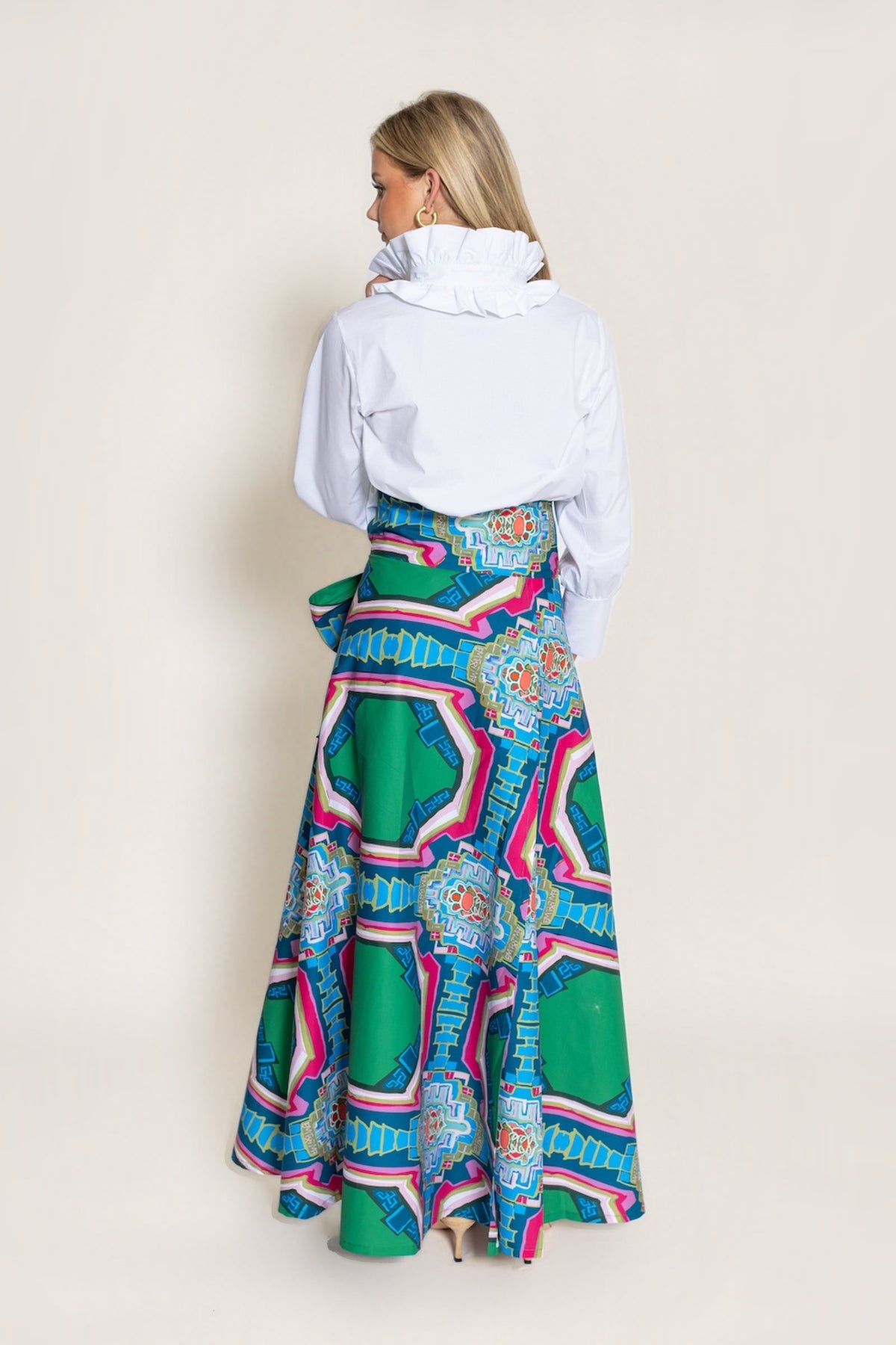A woman shown from the back wearing a white long-sleeve top and a high-waisted, flared maxi skirt with a vibrant green, blue, pink, and purple geometric pattern, set against a solid light beige background.