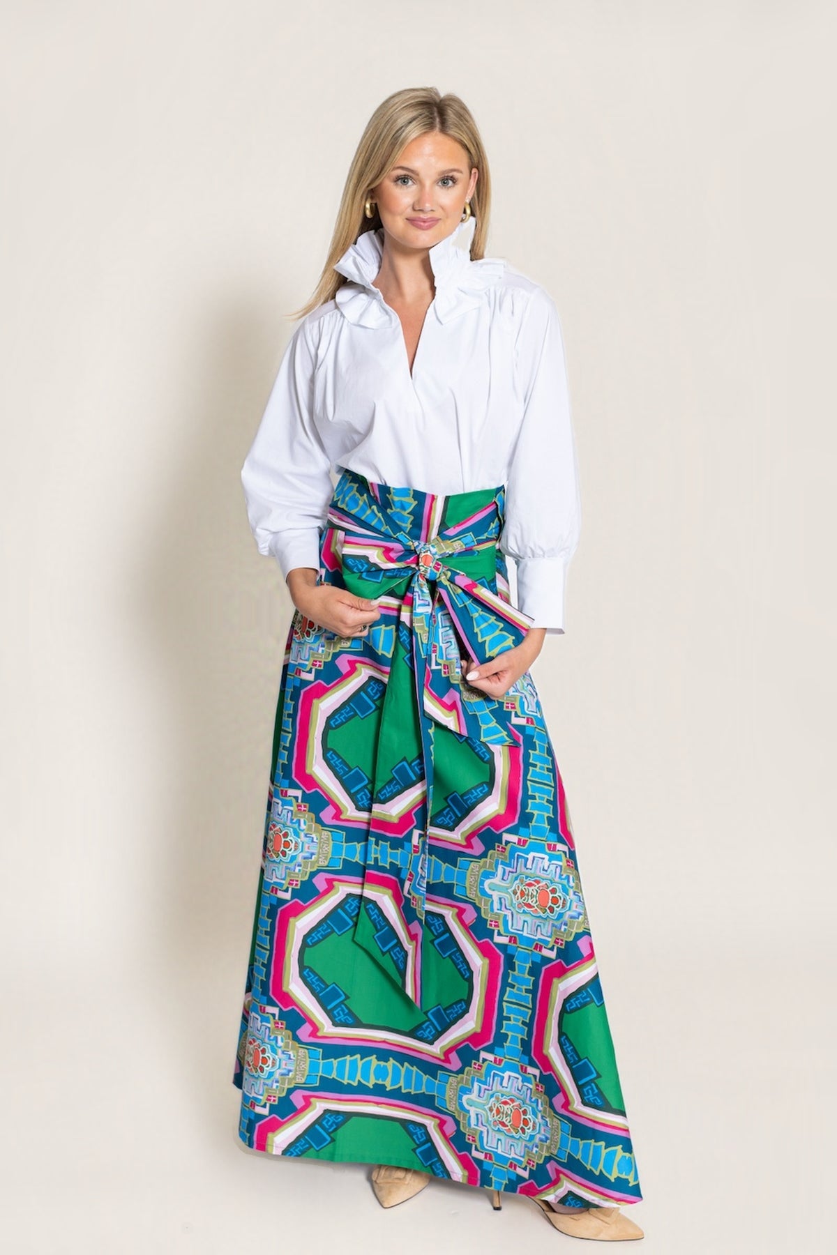 A woman modeling a white long-sleeve collared shirt tucked into a long, wide skirt with a bold geometric pattern in green, blue, pink, and white, standing against a solid light beige background.