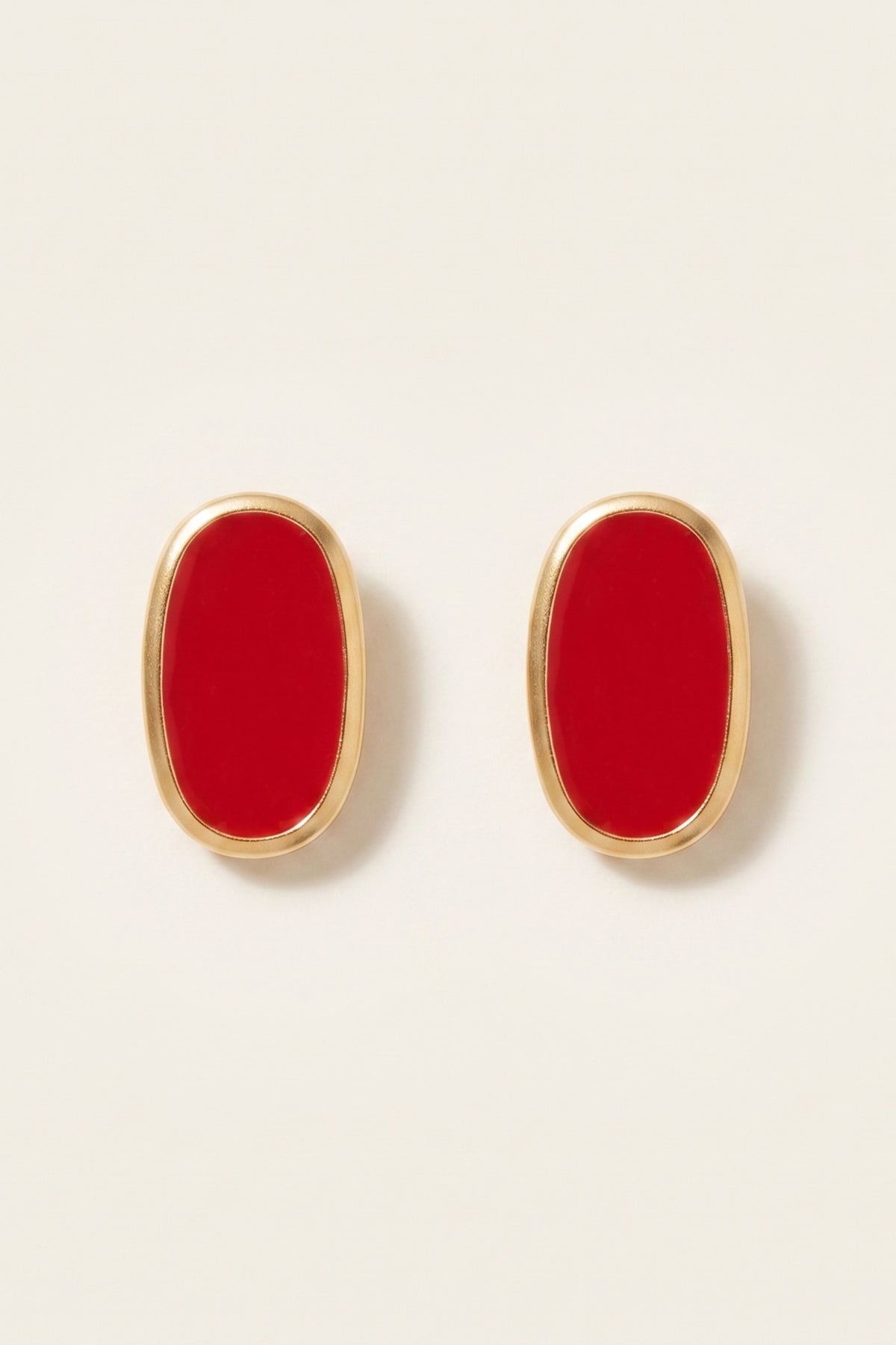 A pair of gold oval stud earrings with vibrant red enamel centers, displayed on a clean, light beige background.