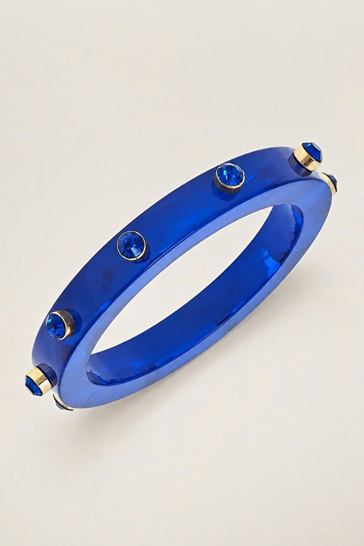 Renee Blue Resin and Rhinestone Bangle, a glossy cobalt blue resin bangle with blue rhinestone accents, on a warm greige backdrop.
