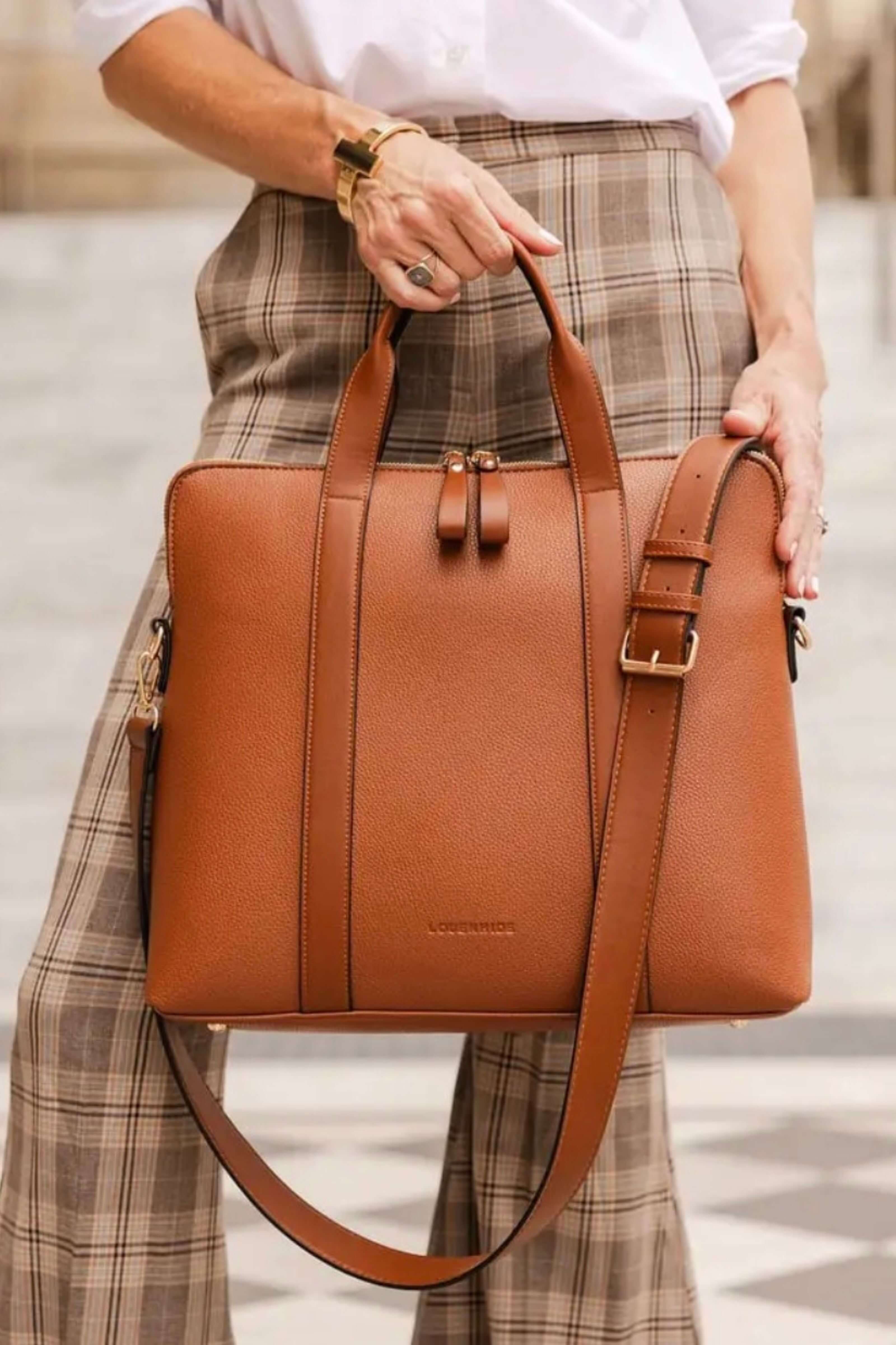 A woman holds a tan leather tote bag with a matching shoulder strap. The bag features a structured rectangular shape, dual top handles, a front zipper closure, and a small embossed brand name near the bottom center.