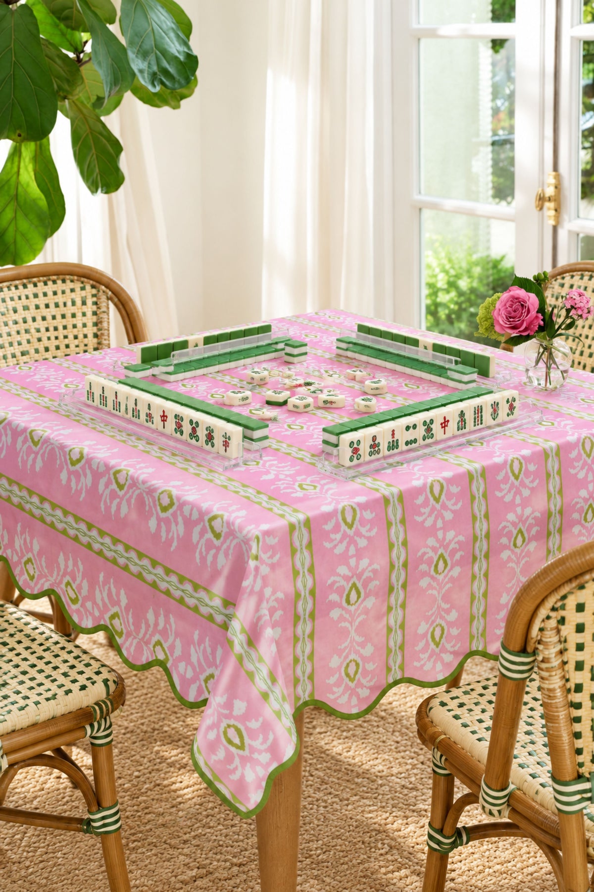 A pink tablecloth with green and white floral patterns draped over a wooden dining table, styled with woven chairs and a mahjong game set in a bright room.