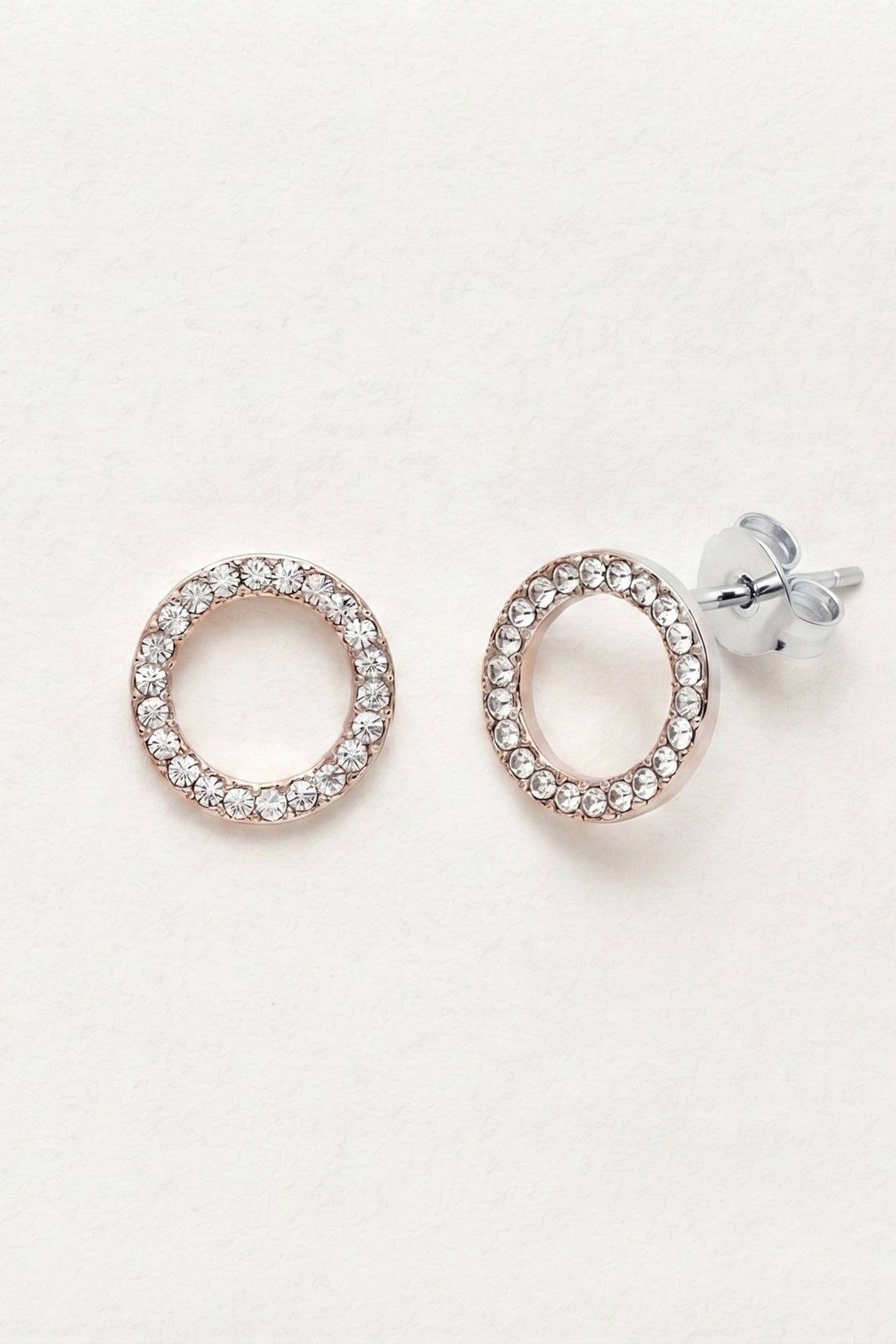 A pair of oval hoop earrings with a single row of clear pavé-set cubic zirconia stones in a rose gold setting, shown on a clean white background. One earring is facing forward, and the other is angled to show the silver-toned post and butterfly back.