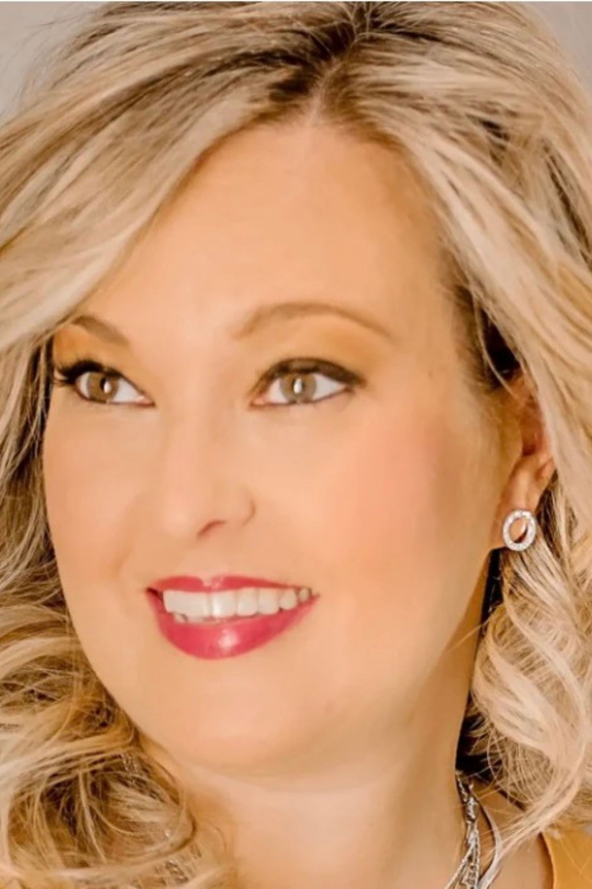 A close-up portrait of a smiling blonde woman wearing a small silver hoop earring, with soft warm lighting and a clean light background.