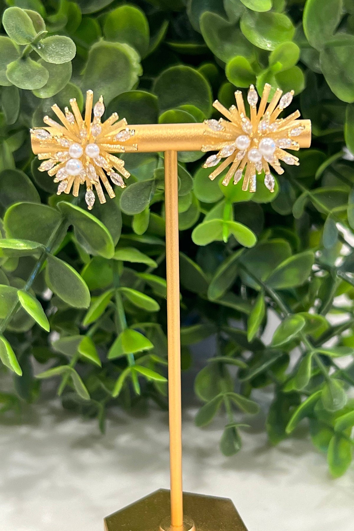 The gold Sunburst earrings with pearls and crystals displayed on a gold T-bar stand in front of greenery.