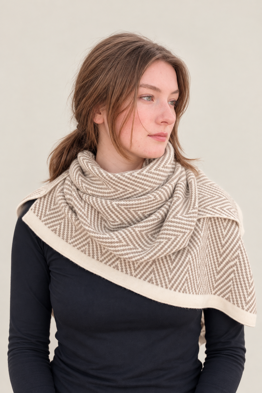 A woman wearing a beige and cream herringbone patterned wrap or scarf draped over her shoulders, paired with a black long-sleeve top, against a solid light beige background.