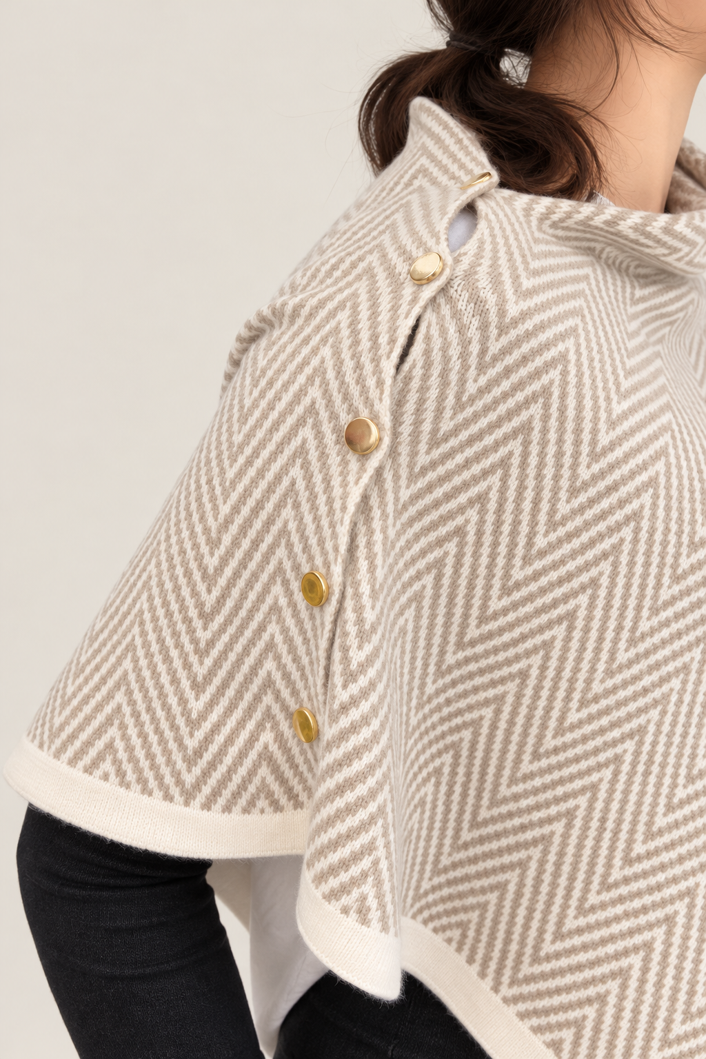 Close-up of a person wearing a beige and white herringbone-patterned cape sweater with gold buttons on the front.