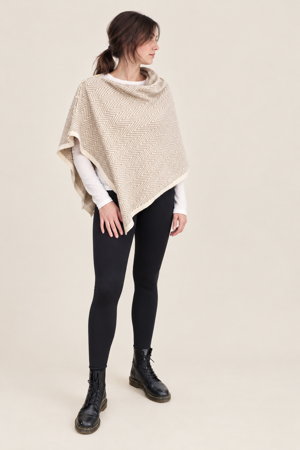 A woman modeling a chunky knit beige poncho over a white long-sleeve top, black leggings, and black lace-up boots, set against a solid light beige background.