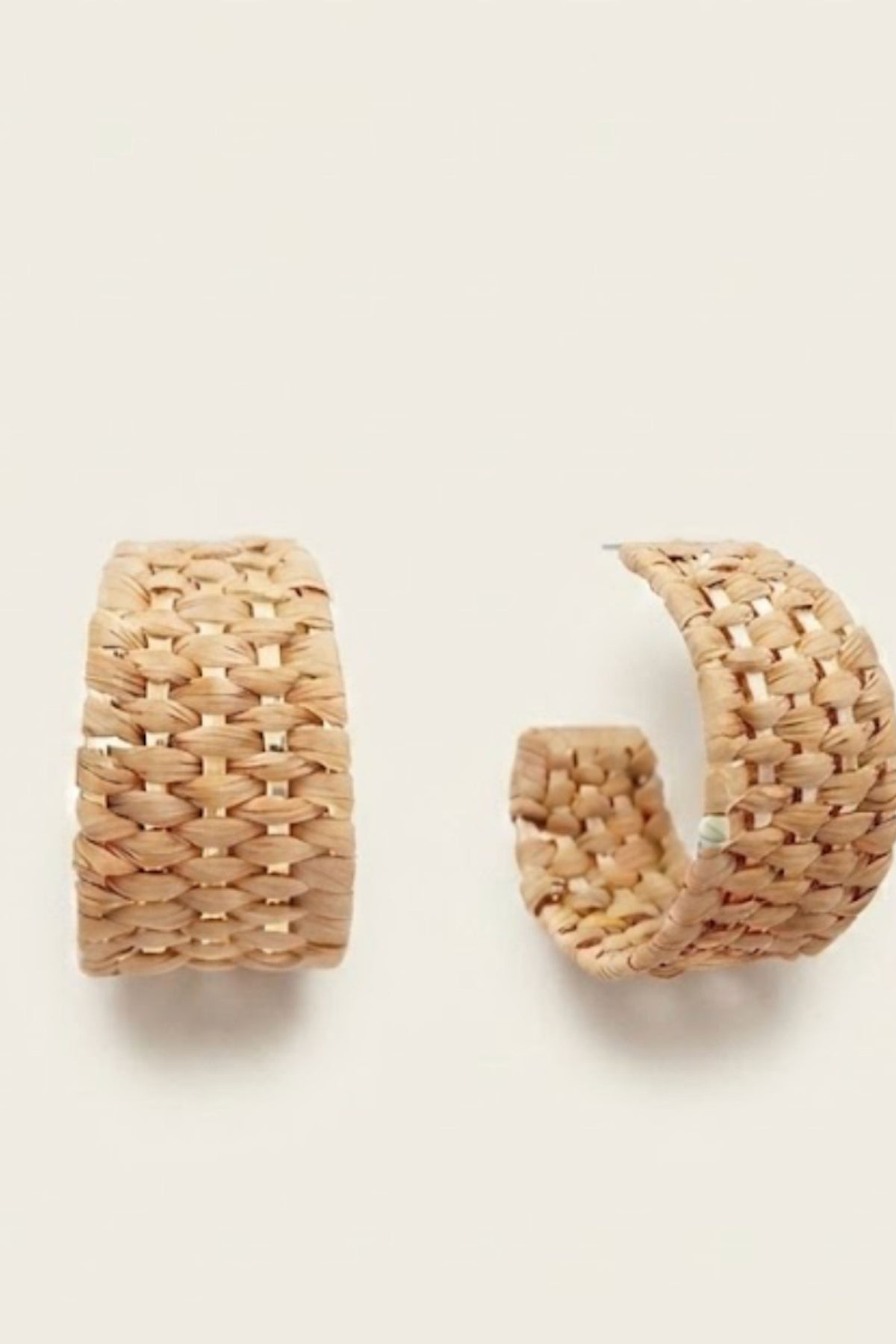 A pair of chunky woven huggie hoop earrings in a natural straw beige color, displayed on a clean off-white background.