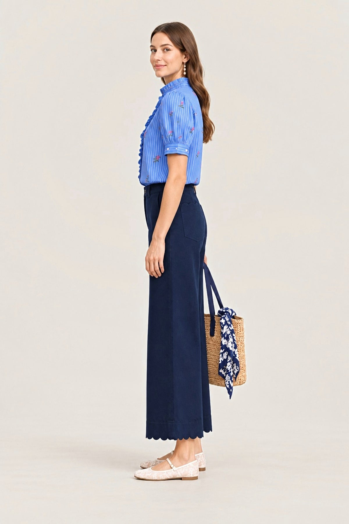 A woman stands in profile wearing a light blue embroidered short-sleeve blouse and a dark navy wide-leg skirt, holding a woven straw tote bag with a blue patterned scarf. She is photographed against a plain off-white studio background.