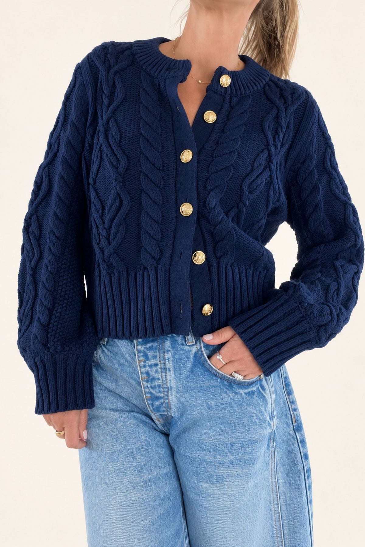 A woman wearing a navy blue cable-knit cardigan with gold buttons, paired with light blue jeans, against a plain cream background.