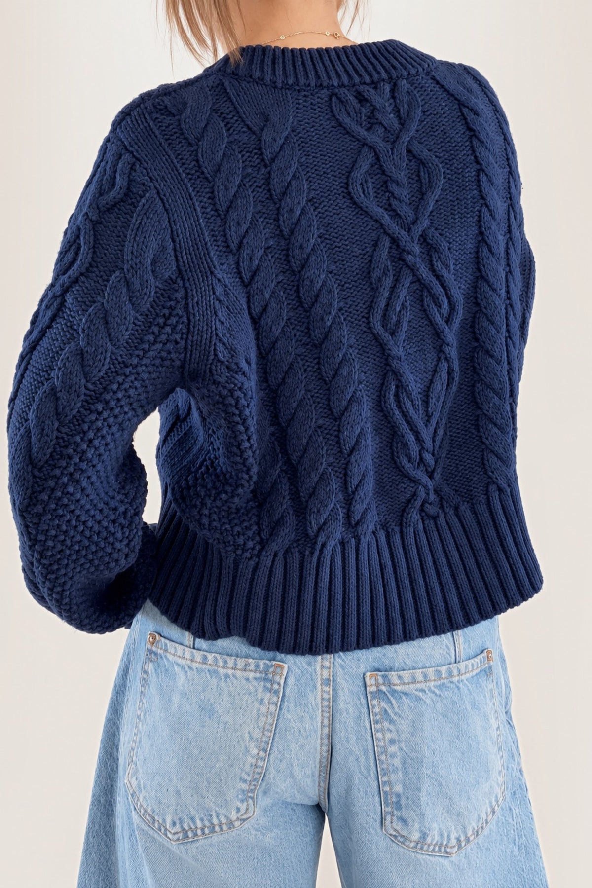 A person shown from the back wearing a navy blue cable-knit sweater and light blue jeans against a plain light background.