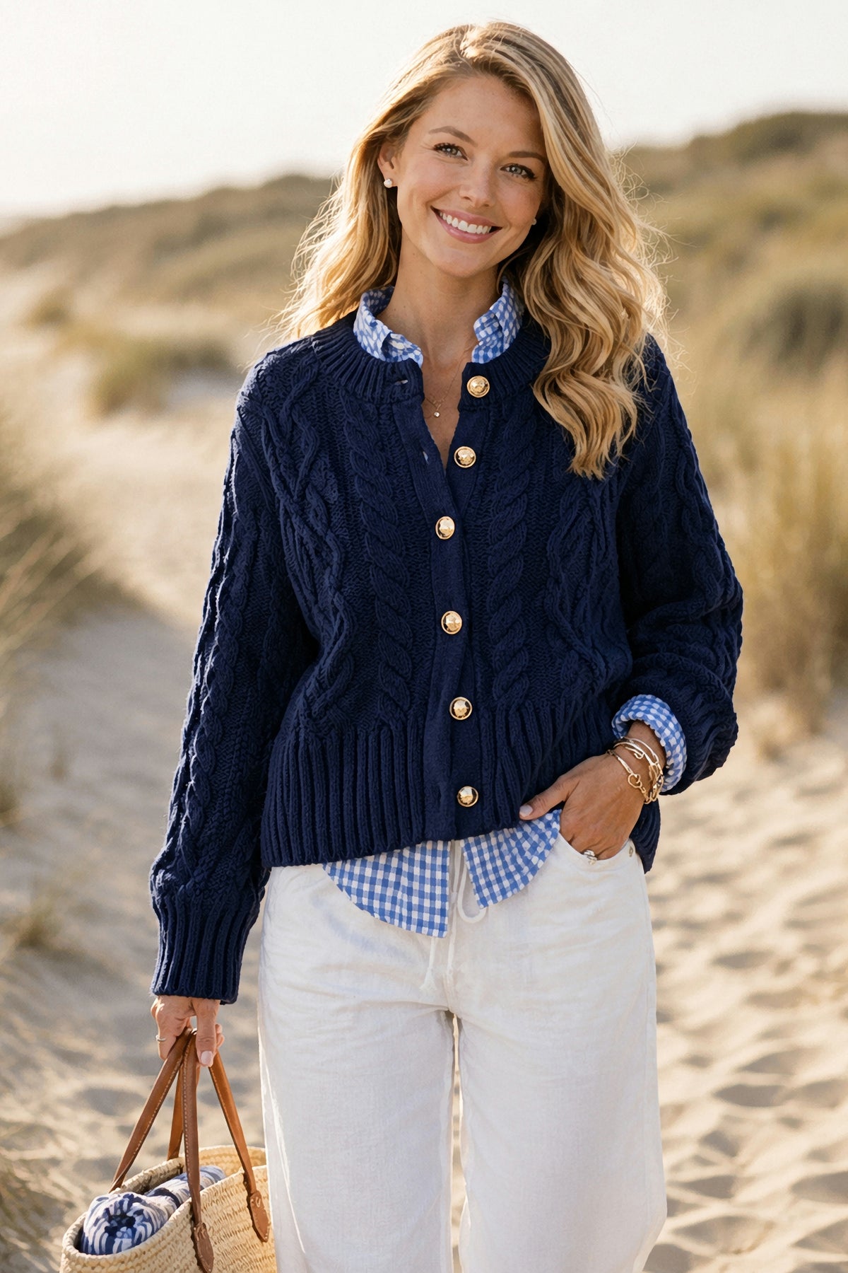 A woman on a sandy beach path wearing The Newport Cable Cardigan in Navy over a gingham shirt with white jeans