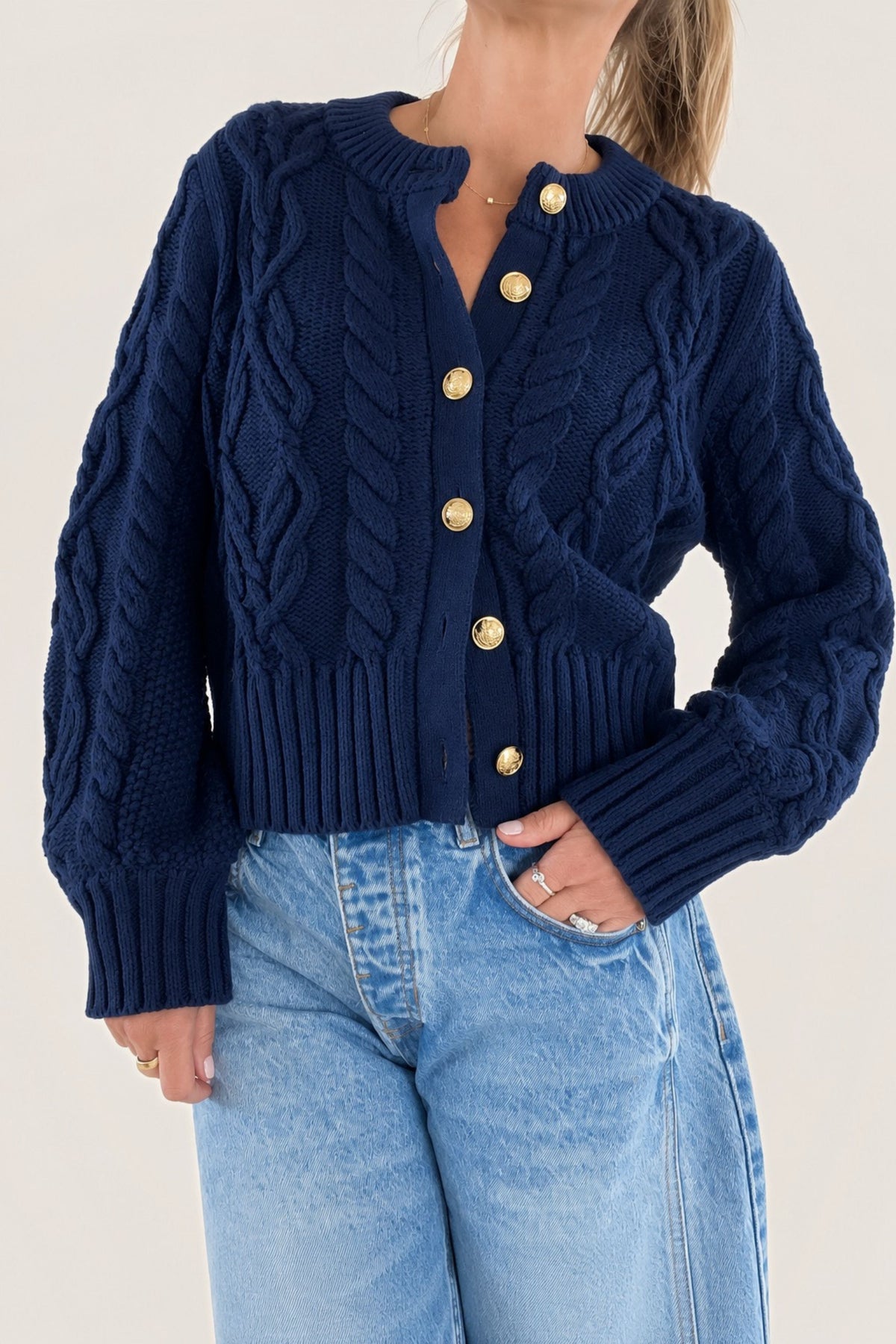A woman wearing a navy blue cable-knit cardigan with gold buttons, paired with light blue jeans, against a plain cream background.