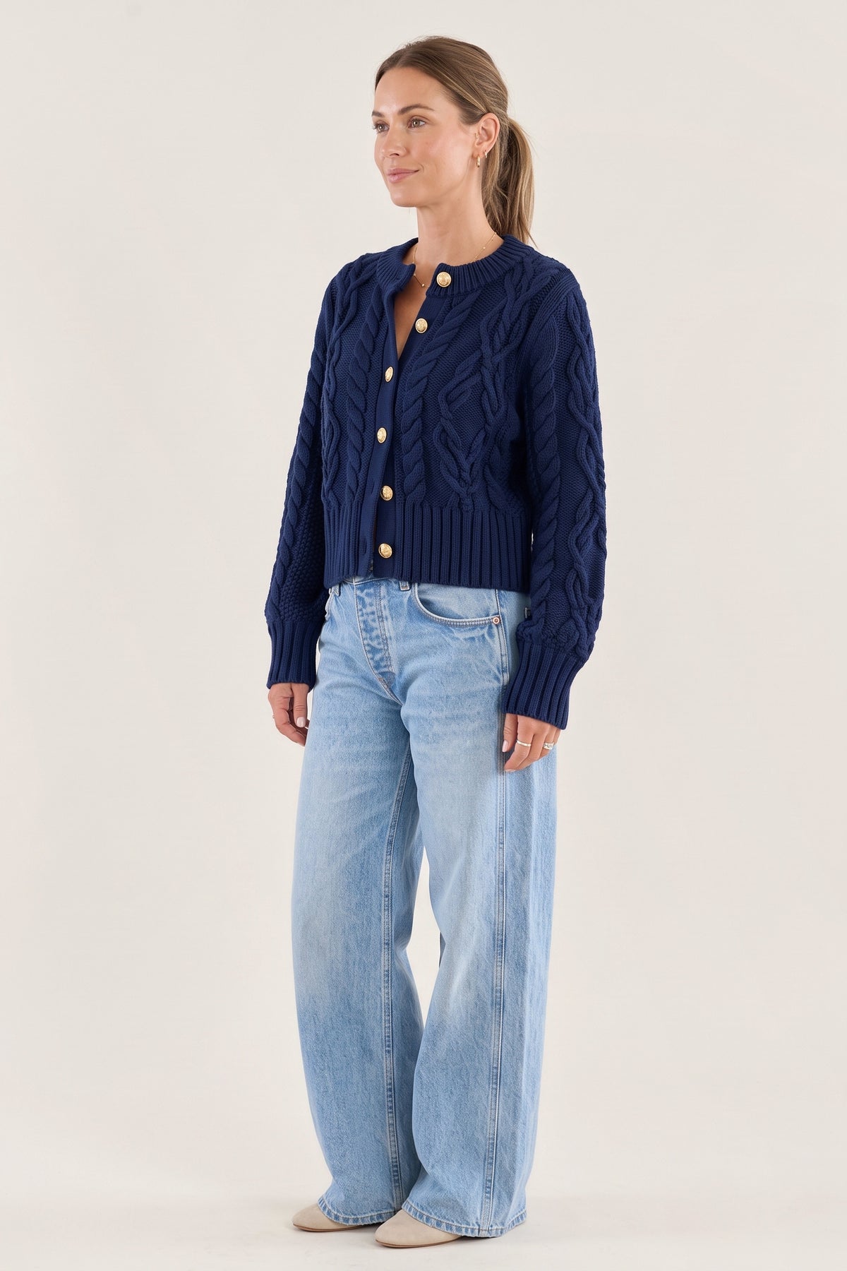 A woman in a three-quarter view wearing The Newport Cable Cardigan in Navy, buttoned, with light wash wide-leg jeans against a plain studio backdrop