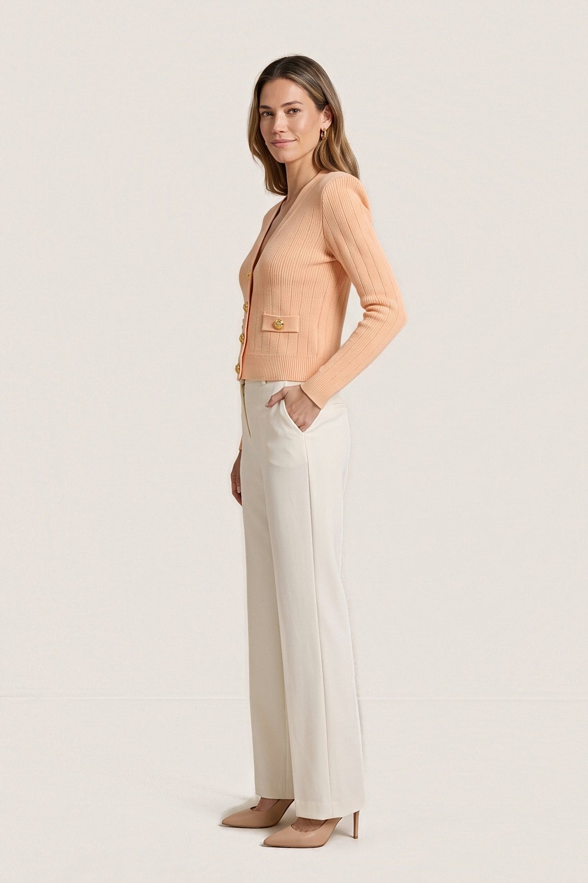 Side view of the peach Timeless Knit Cardigan showing the structured shoulder, fitted long sleeve, button-tab pocket and cropped length at the waist.