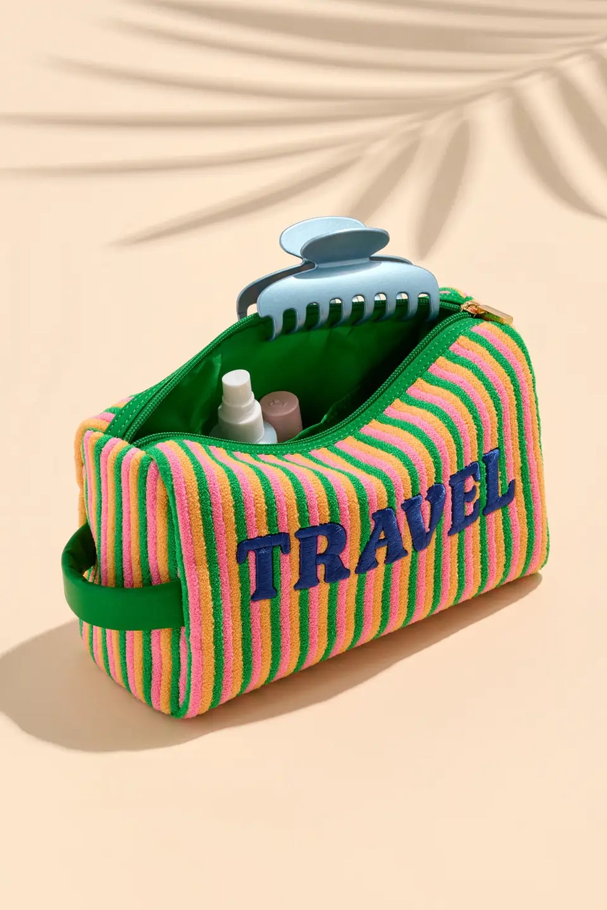 The Travel zip pouch open on a peach background, showing its green lining with a hair clip and toiletries inside.