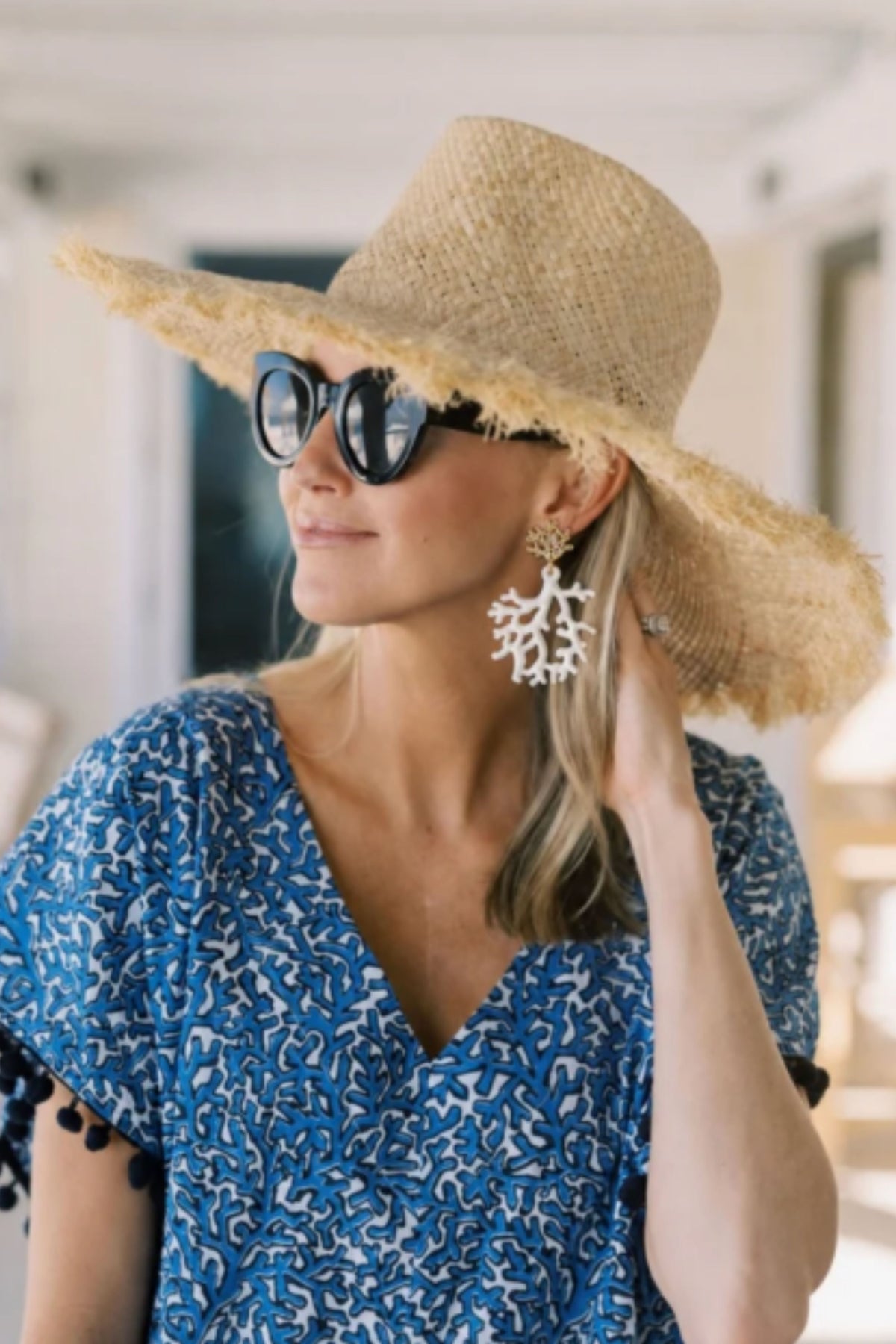 A woman wearing a wide-brimmed straw hat, black sunglasses, and a blue and white patterned top, with a large white coral-shaped earring visible on one ear.