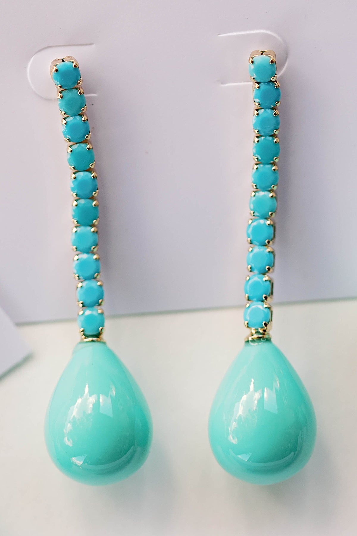 The turquoise Swingy statement earrings hanging from a display card, showing the line of turquoise stones set in gold and the teardrop.