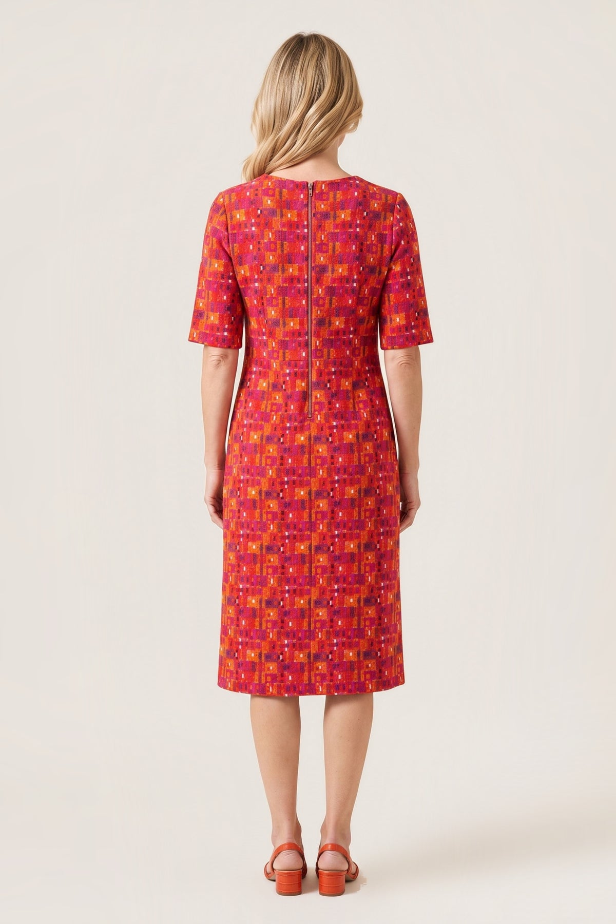 A woman viewed from the back wearing a knee-length short-sleeve dress with a dense geometric pattern in red, orange, yellow, and purple, paired with brown sandals, against a solid light beige background.