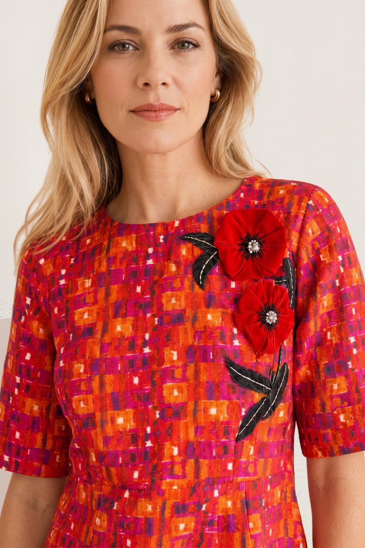 A woman wearing a short-sleeve dress with a vibrant orange, red, and magenta geometric pattern, featuring large red poppy appliques with black leaves on the chest.