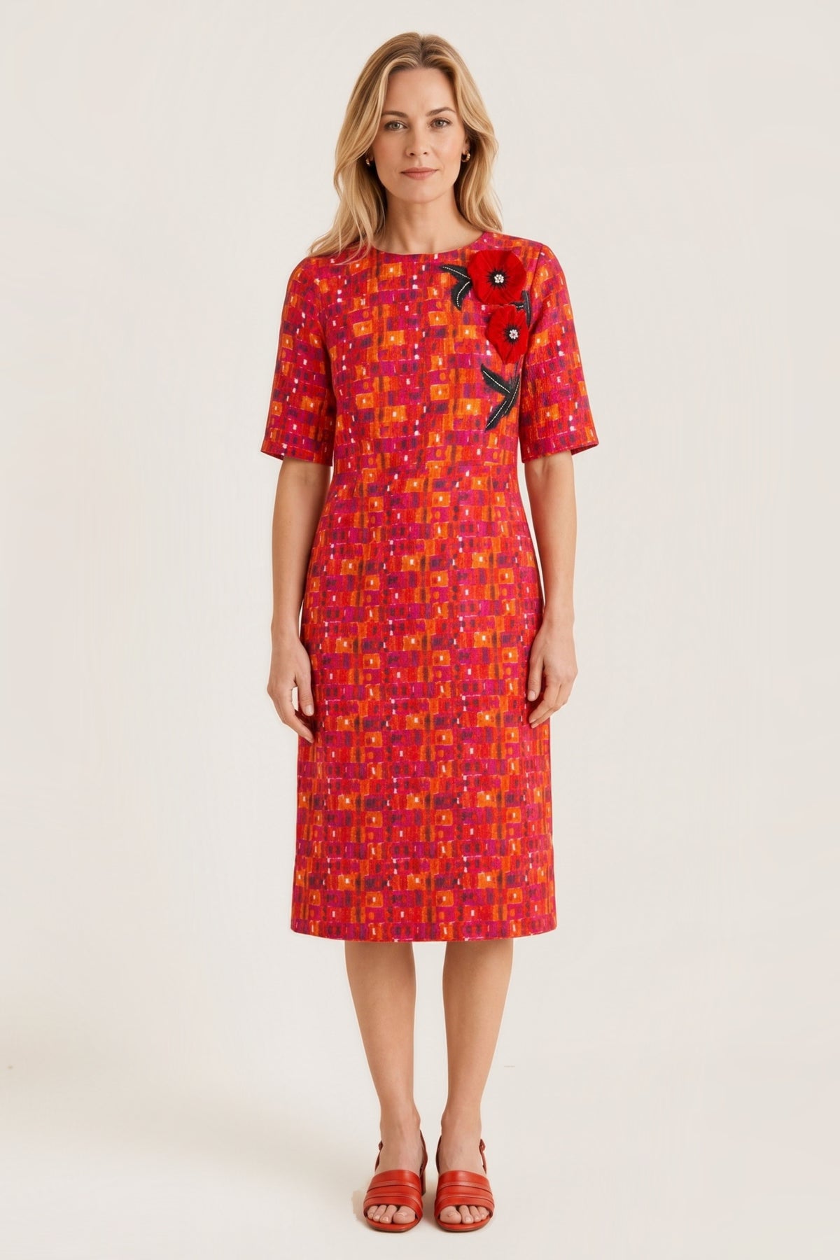 A woman modeling a short-sleeved knee-length dress with a vibrant red, orange, and yellow geometric pattern, featuring a large red rose applique on the left chest, set against a plain beige studio background.