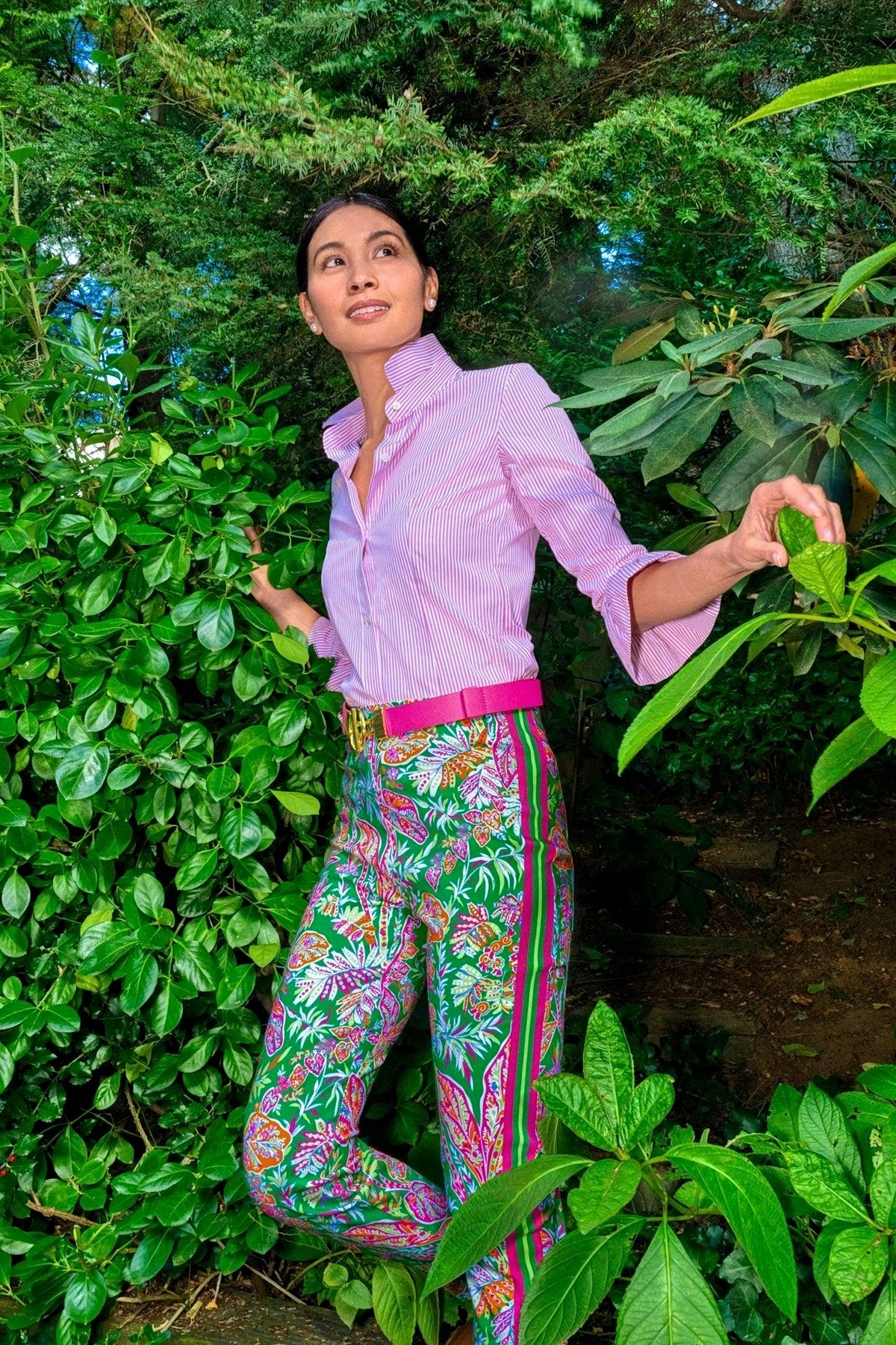 A woman stands among green foliage in a pink striped shirt, pink belt and kelly green vine print pull-on pants.