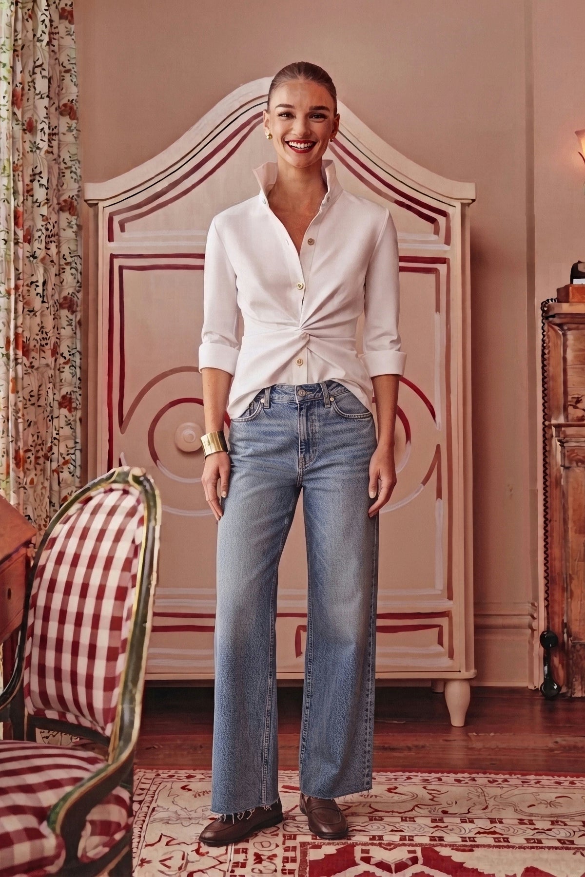 A woman stands in a vintage-style bedroom setting wearing a white long-sleeve button-up shirt tied at the waist and light-wash blue wide-leg jeans.