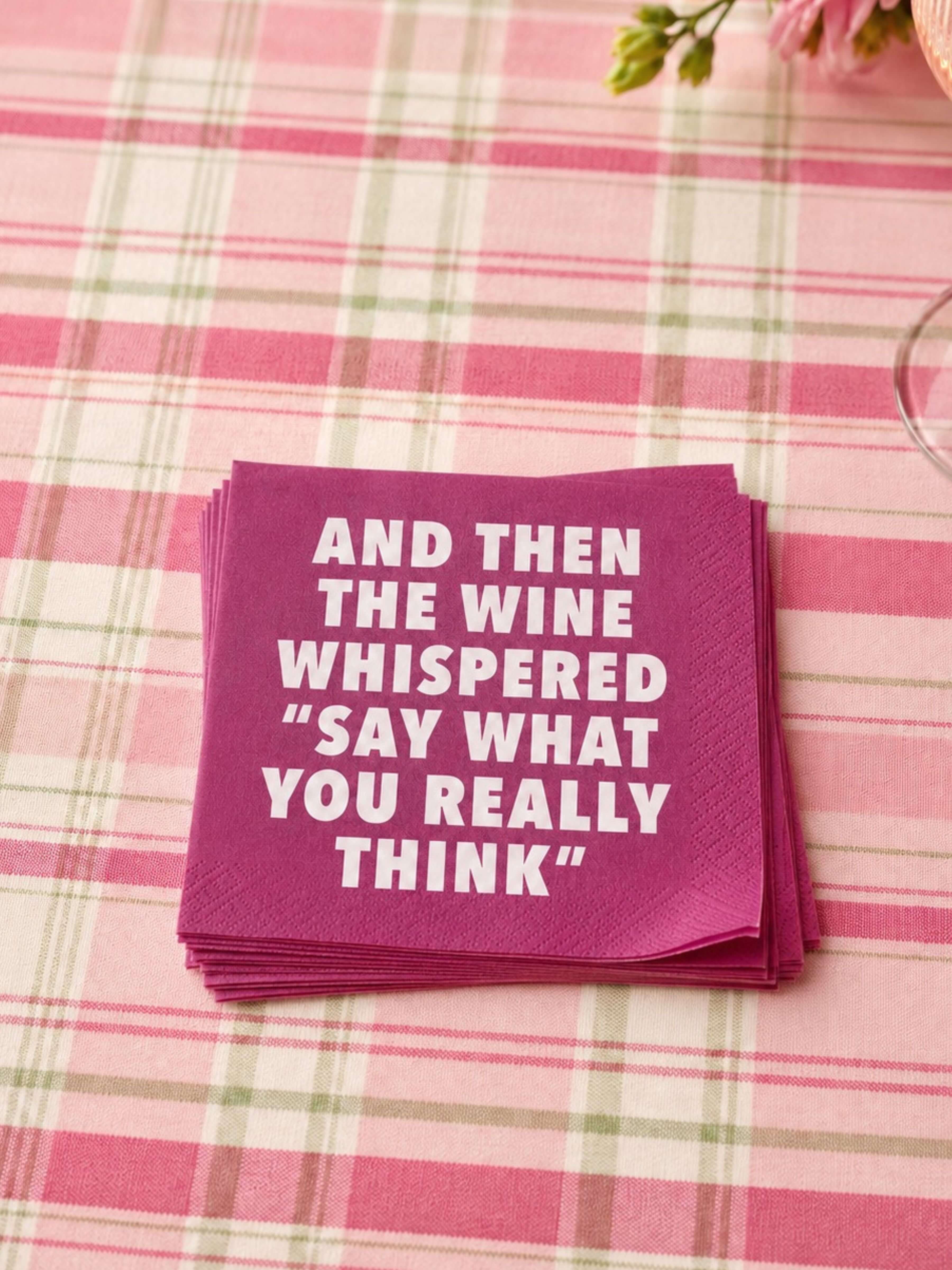A stack of magenta paper napkins with bold white text reading 'AND THEN THE WINE WHISPERED "SAY WHAT YOU REALLY THINK"' placed on a pink and white plaid tablecloth.